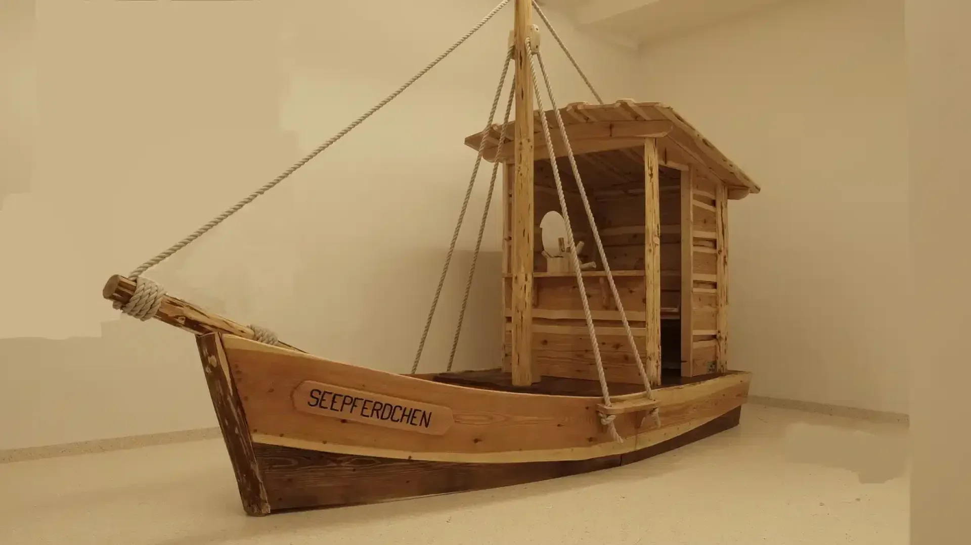 A wooden play boat with a small cabin and mast is set indoors on a smooth, light-colored floor. The word SEEPFERDCHEN is written on the side of the boat.