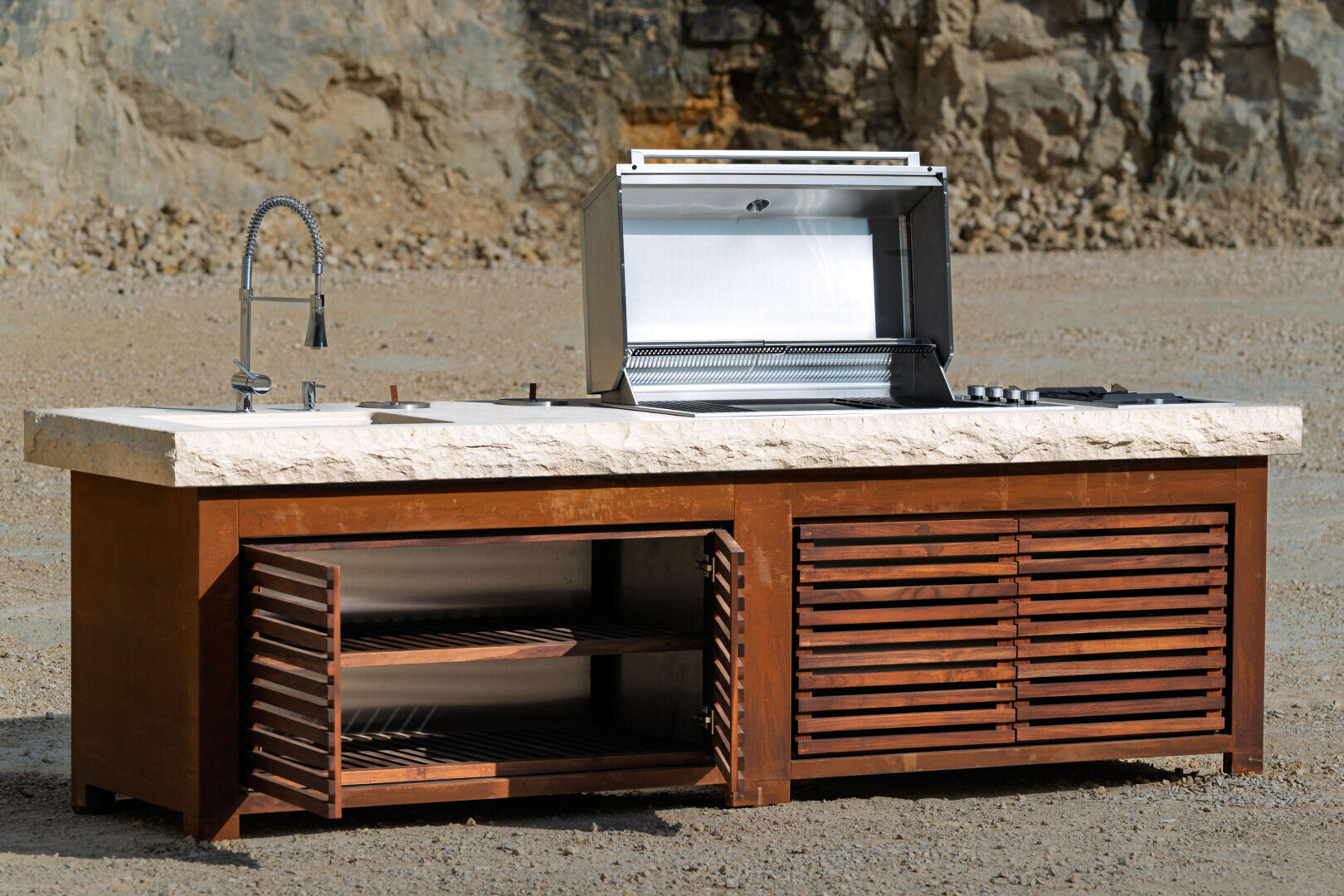 Eine moderne Außenküche mit eingebautem Grill, offenem Edelstahldeckel, Spüle mit geschwungenem Wasserhahn und Holzlattenschränken auf einer Kiesfläche mit felsigem Hintergrund.