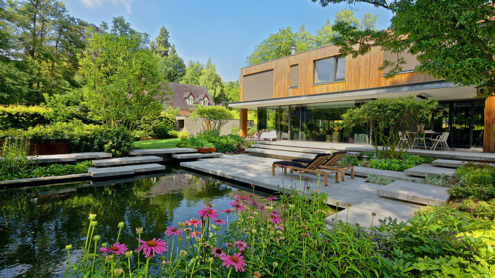 Modernes Haus mit großen Fenstern und Blick auf einen Landschaftsgarten mit einem Teich mit Betontrittsteinen, blühenden Blumen, Liegestühlen und üppigem Grün unter einem klaren blauen Himmel.