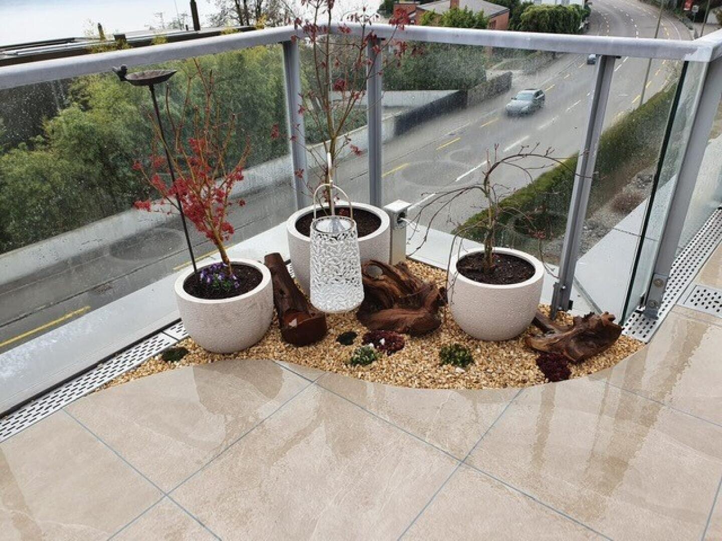 Drei Topfpflanzen und eine weiße dekorative Laterne stehen auf einem Kiesbett neben Treibholzstücken auf einem Balkon mit Glasgeländer und Blick auf eine Straße und Bäume.