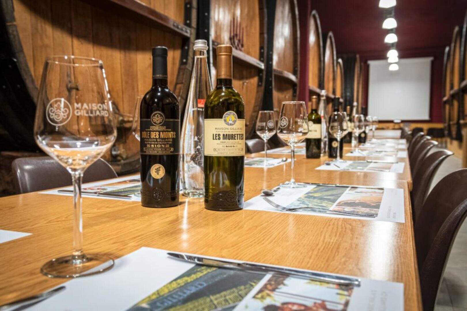 Ein langer Holztisch, der für eine Weinprobe gedeckt ist, mit Weinflaschen, Gläsern, Wasserflaschen und Tischsets, die fein säuberlich angeordnet sind, umgeben von Holzfässern in einem warm beleuchteten Keller.