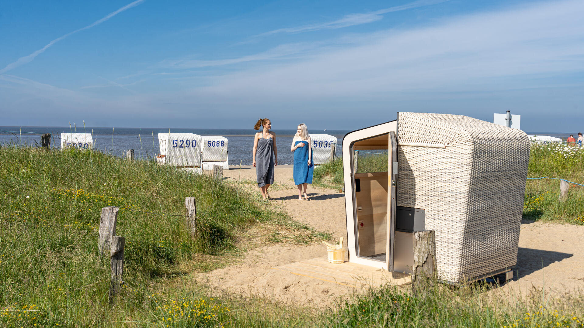 Zwei Frauen gehen auf einem sandigen Weg in Richtung Strand, vorbei an weißen Strandkörben mit Nummern. Im Vordergrund steht eine Strandhütte aus Weidengeflecht, und grasbewachsene Dünen säumen die Seiten. Der Himmel ist blau und leicht bewölkt.