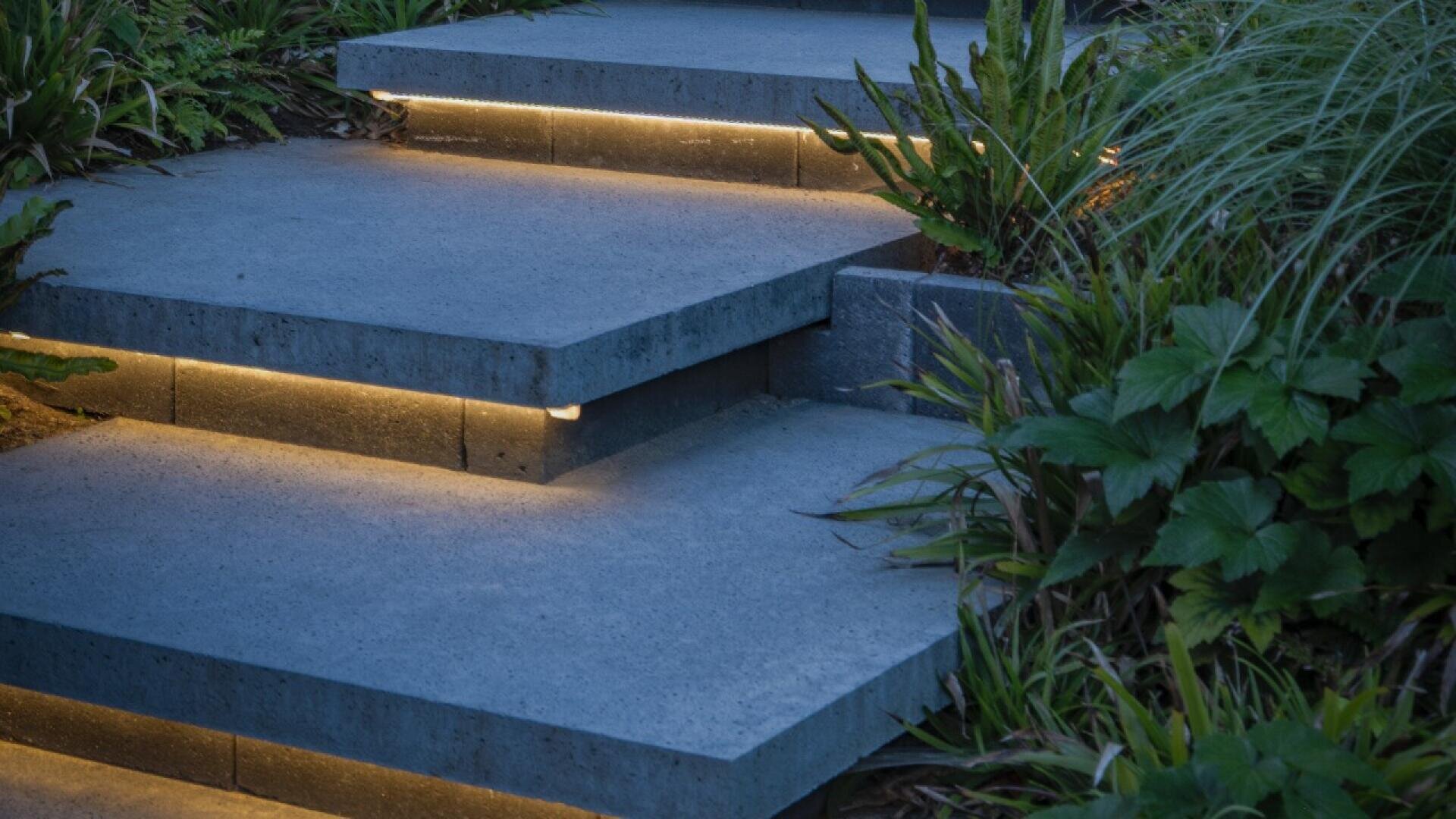 Concrete steps with built-in LED strip lighting underneath each step, surrounded by green plants and foliage, creating a modern and illuminated outdoor path.
