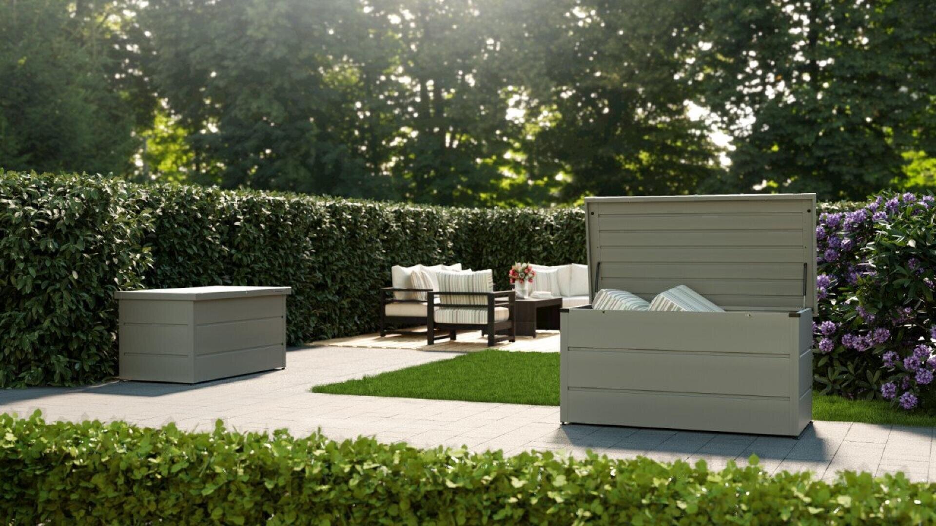 Outdoor patio with a cushioned seating area, coffee table, and two large, light gray storage boxes—one open and filled with pillows—surrounded by green hedges and flowering bushes on a tiled surface.