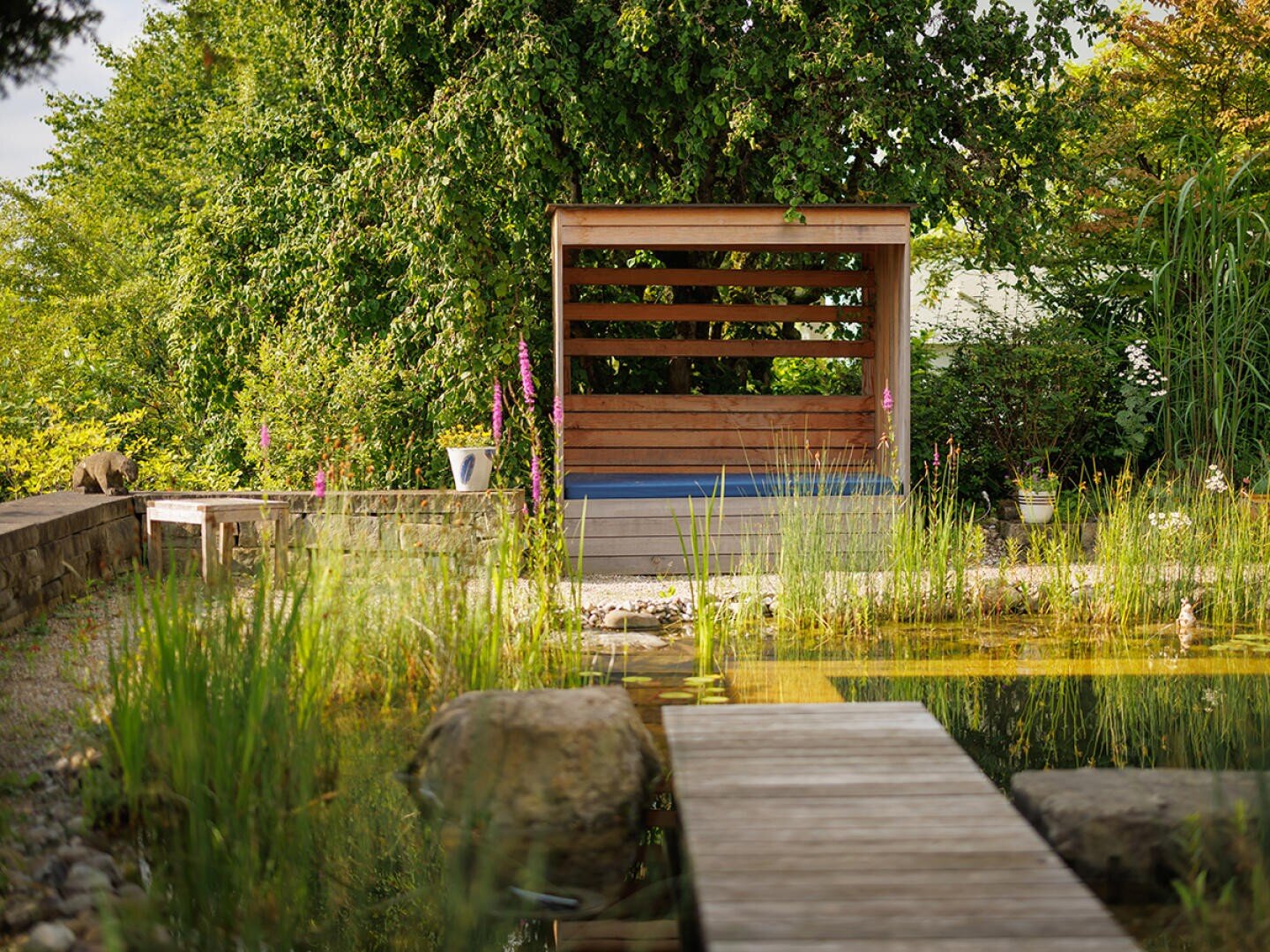 Eine überdachte Holzbank steht an einem Teich, der von hohem Gras, Steinen und blühenden Blumen umgeben ist. Ein Holzsteg führt über den Teich zur Bank, die von üppig grünen Bäumen beschattet wird.