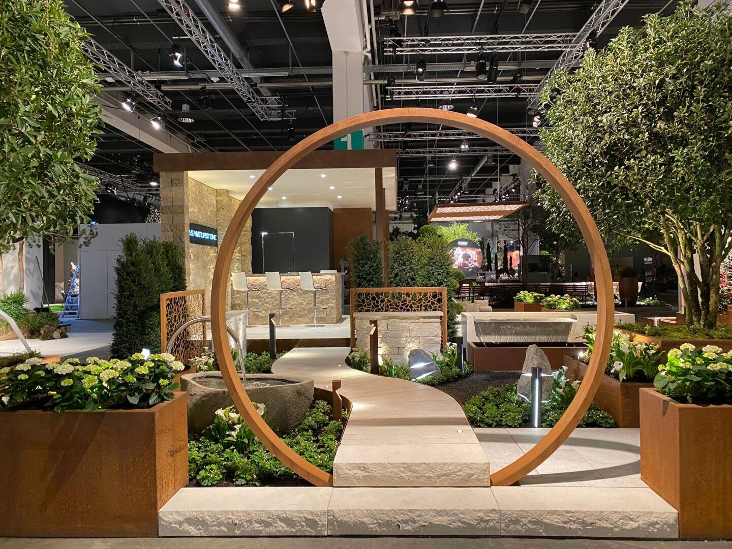 A modern indoor garden features a large round metal arch over a stone path, surrounded by lush greenery, flowers and fountains, with trees and architectural elements in the background.