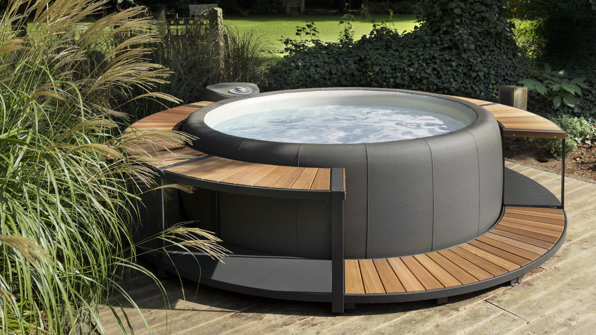 Ein runder, moderner Whirlpool im Freien mit sprudelndem Wasser ist von einer Holzterrasse umgeben. Er steht auf einer Holzterrasse mit üppigen Grünpflanzen und einem grasbewachsenen Hof im Hintergrund.