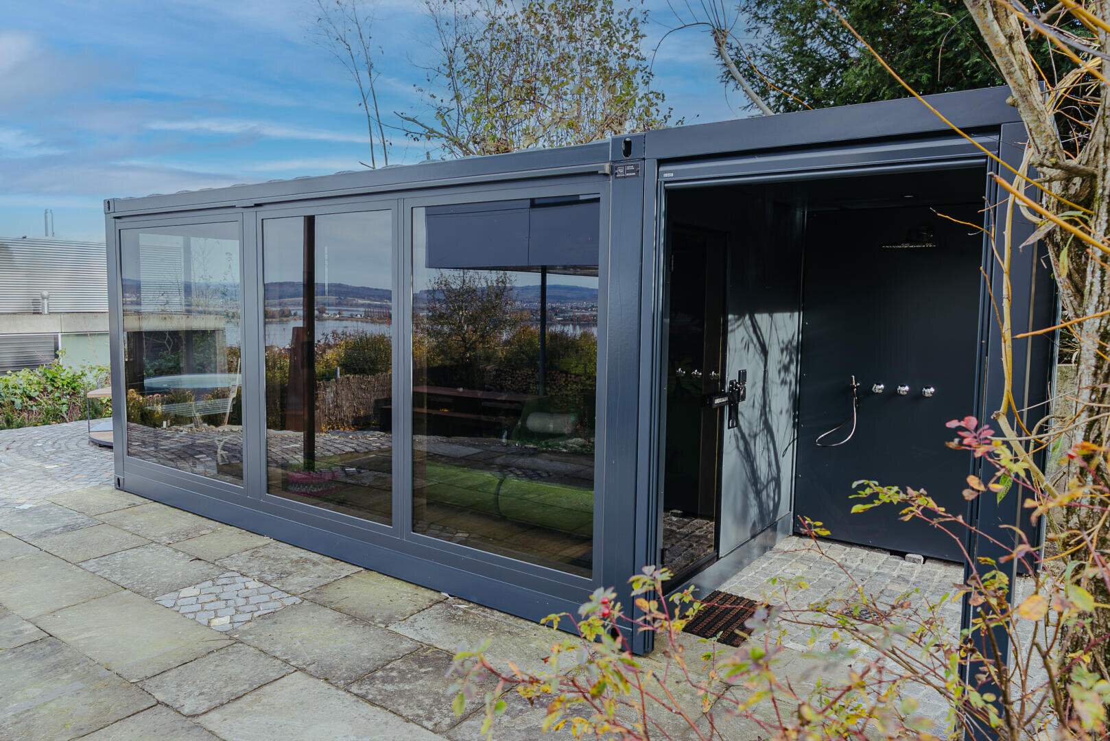 Ein modernes Gartenzimmer mit großen Glastüren und Fenstern steht auf einer Steinterrasse, umgeben von Pflanzen. Auf einer Seite ist eine Außendusche zu sehen, und im Hintergrund stehen Bäume unter einem teilweise bewölkten Himmel.