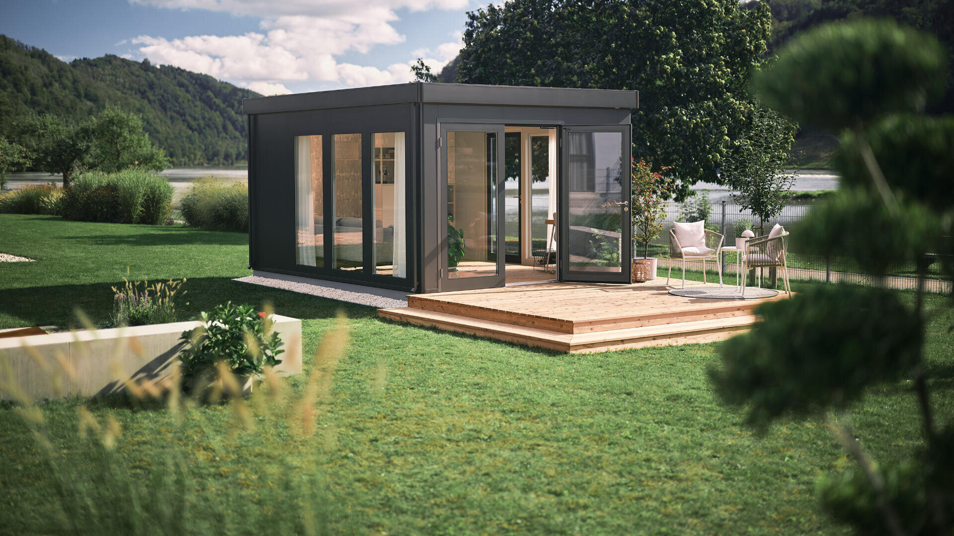 Ein modernes, kleines Glashaus steht auf einer Holzterrasse in einem grünen Garten mit Bäumen, Terrassenmöbeln und einem See und Hügeln im Hintergrund unter einem teilweise bewölkten Himmel.