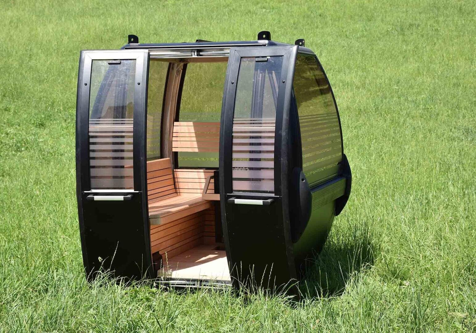 A black and glass gondola cabin with wooden seating sits on green grass in an open field, detached from any cable or support structure.