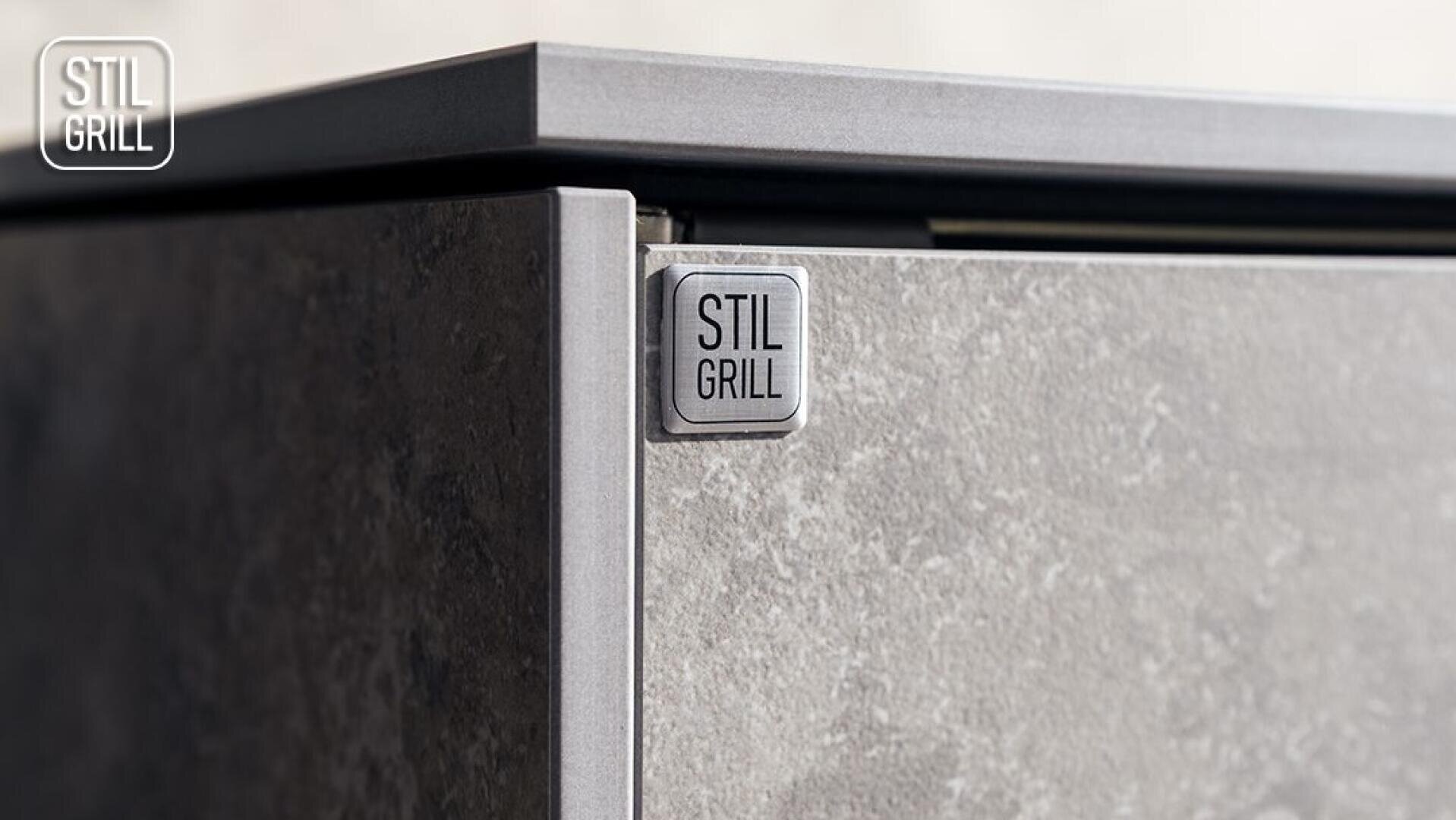 Nahaufnahme der Ecke eines grauen Geräts mit Steinstruktur und einer Metallplakette mit der Aufschrift STIL GRILL auf der Vorderseite. Das STIL GRILL-Logo ist ebenfalls in der oberen linken Ecke des Bildes zu sehen.