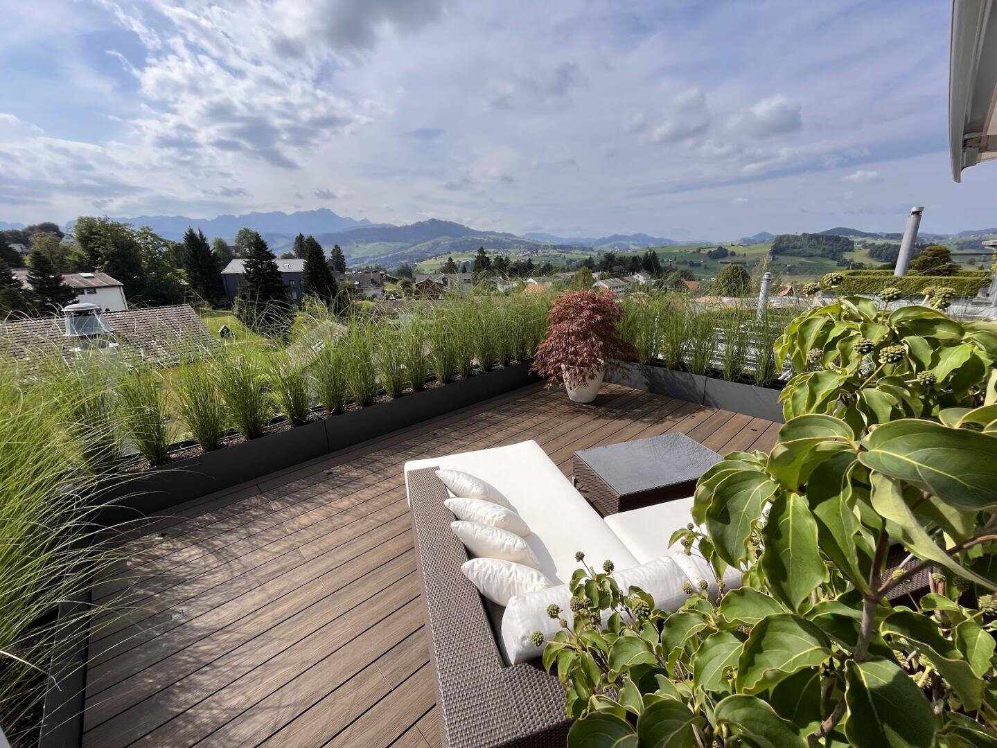 Eine moderne Dachterrasse mit Liegestühlen, dekorativen Kissen, Topfpflanzen und üppigem Grün bietet einen Blick auf sanfte Hügel, Häuser und ferne Berge unter einem teilweise bewölkten Himmel.