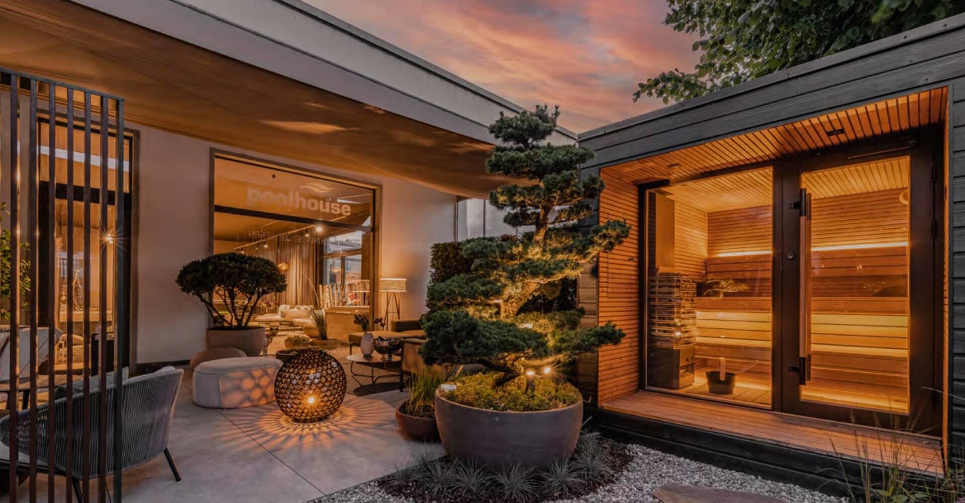 A modern outdoor patio at sunset, featuring a cozy seating area, potted plants, warm lighting, and a wooden sauna room with glass doors.