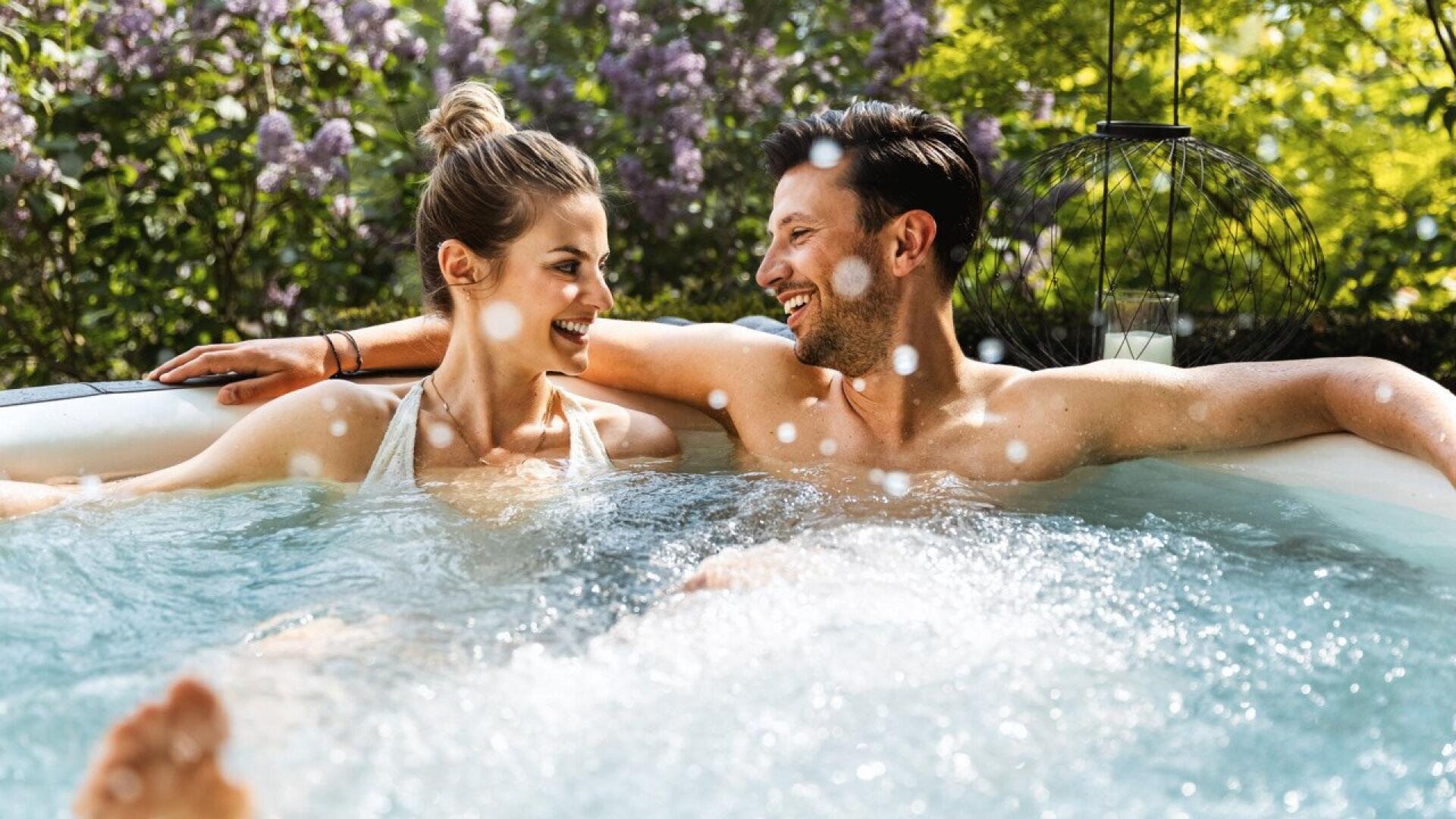 A smiling couple relaxes together in an outdoor hot tub, surrounded by greenery and blooming lilac bushes, enjoying a sunny day.