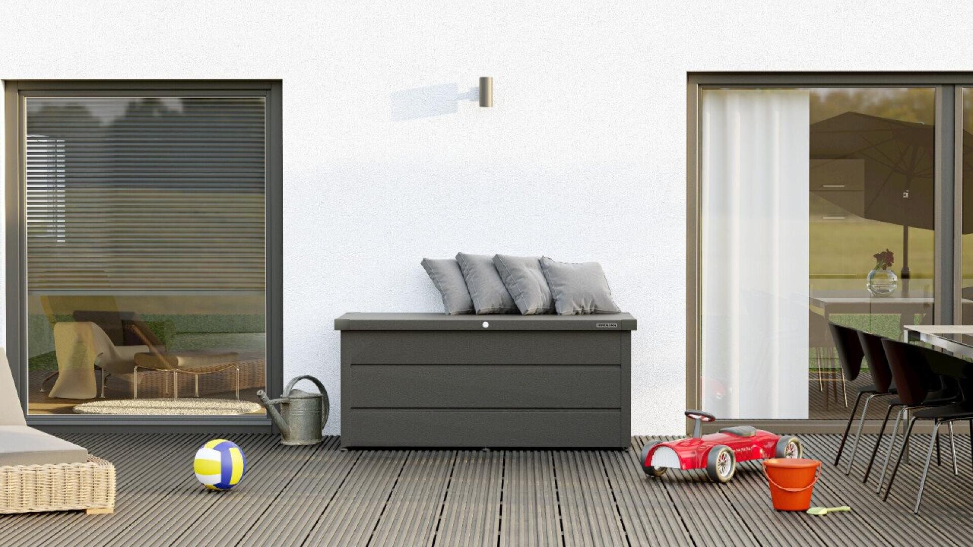 A modern patio with a storage box topped with gray cushions, children’s toys (ball, red ride-on car, orange bucket), a watering can, table and chairs, and glass doors reflecting outdoor furniture and greenery.