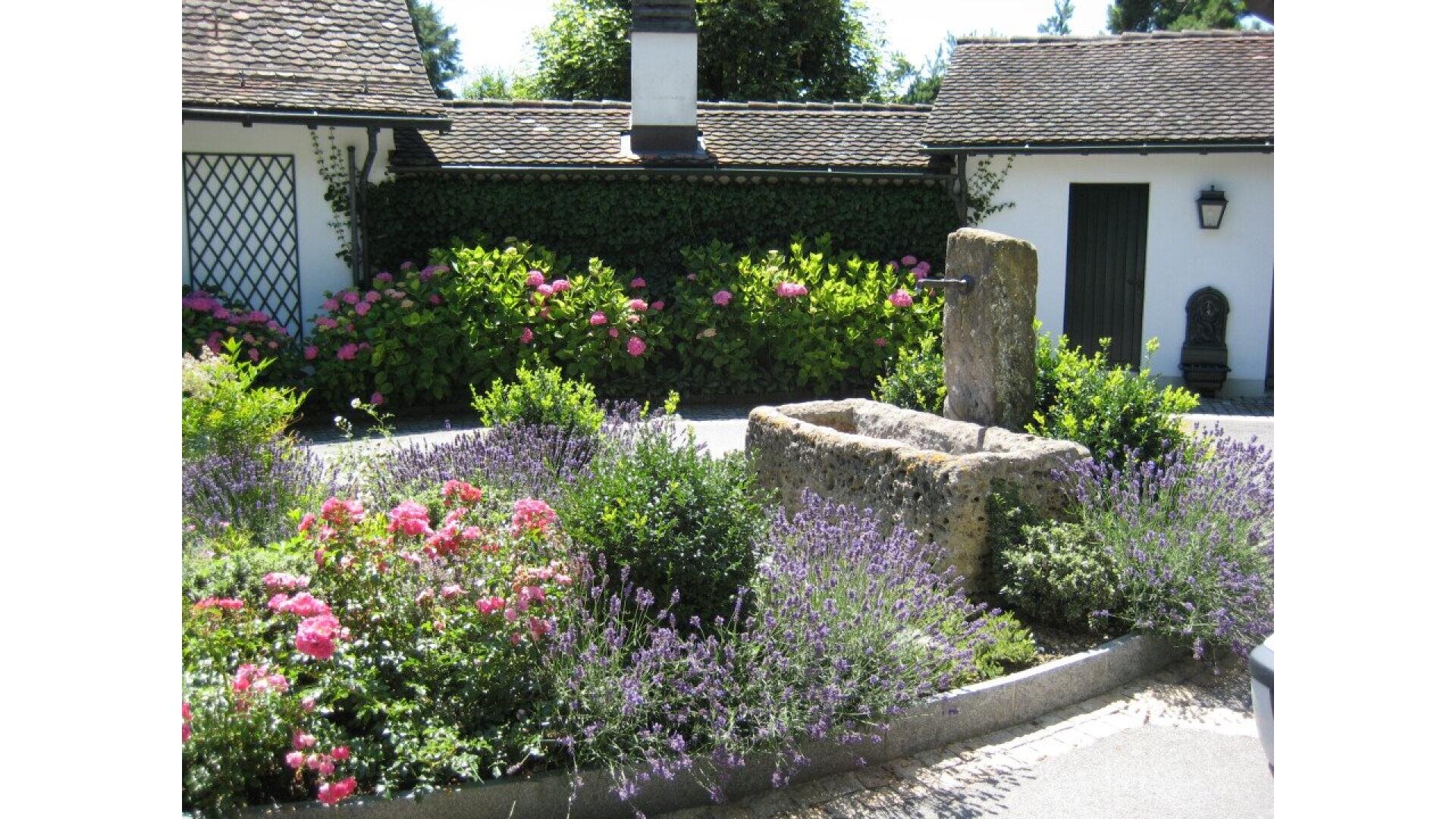 A stone water fountain surrounded by lavender, pink flowers, and greenery in front of a white building with a tiled roof and wooden doors on a sunny day.