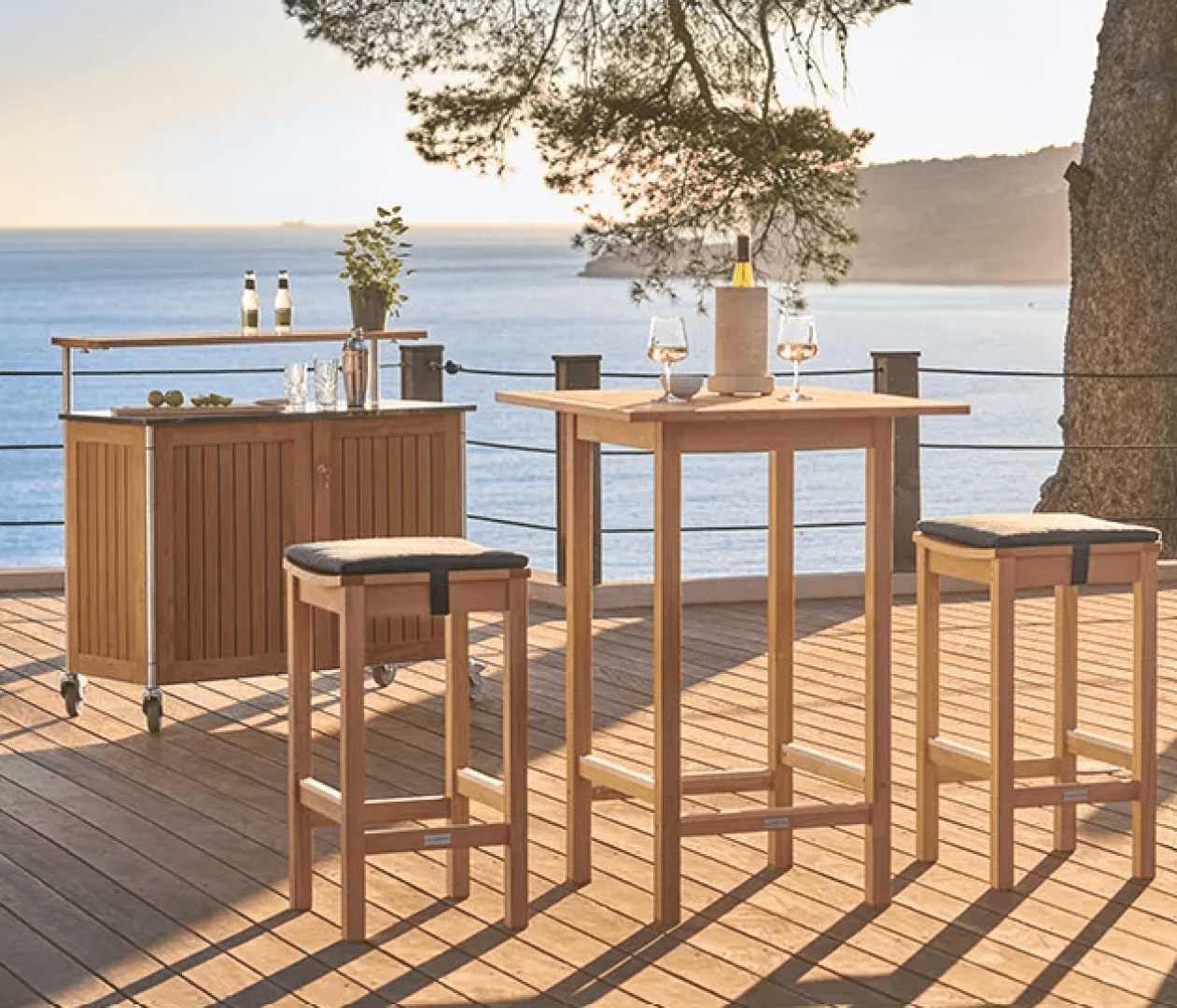 Outdoor wooden bar table with two stools and a serving cart on a deck overlooking the ocean, with two wine glasses, a bottle, and sunlight filtering through trees.
