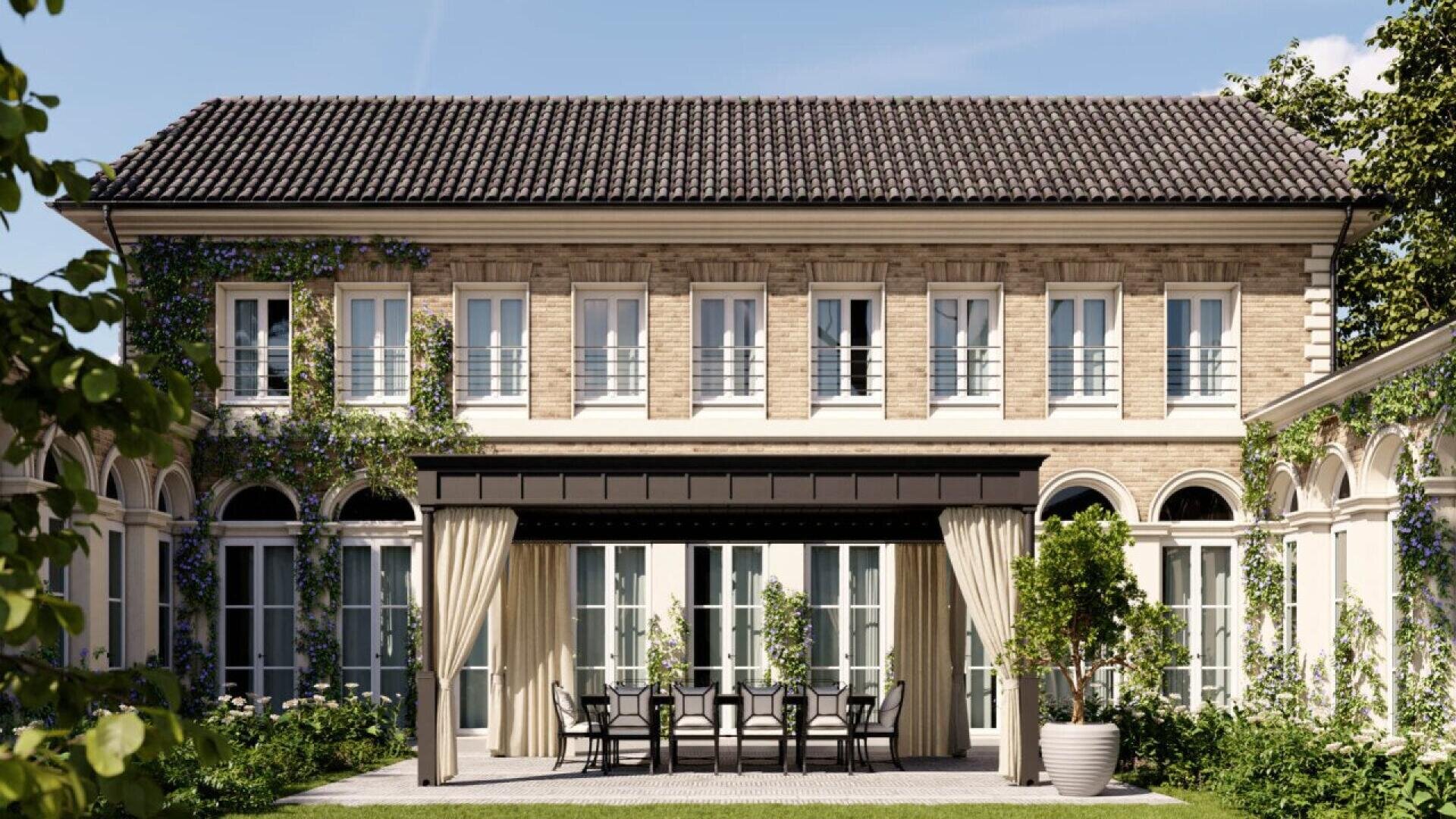 A two-story house with a tiled roof, beige brick walls, and multiple white-framed windows. There is a patio with outdoor dining furniture, potted plants, greenery, and draped curtains under a metal pergola.