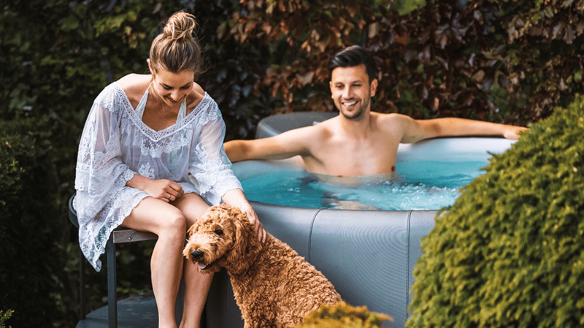 Eine Frau sitzt neben einem Whirlpool und streichelt einen lockigen Hund, während sich ein Mann lächelnd im Whirlpool entspannt. Üppiges Grün und Sträucher umgeben sie in einer Außenanlage.