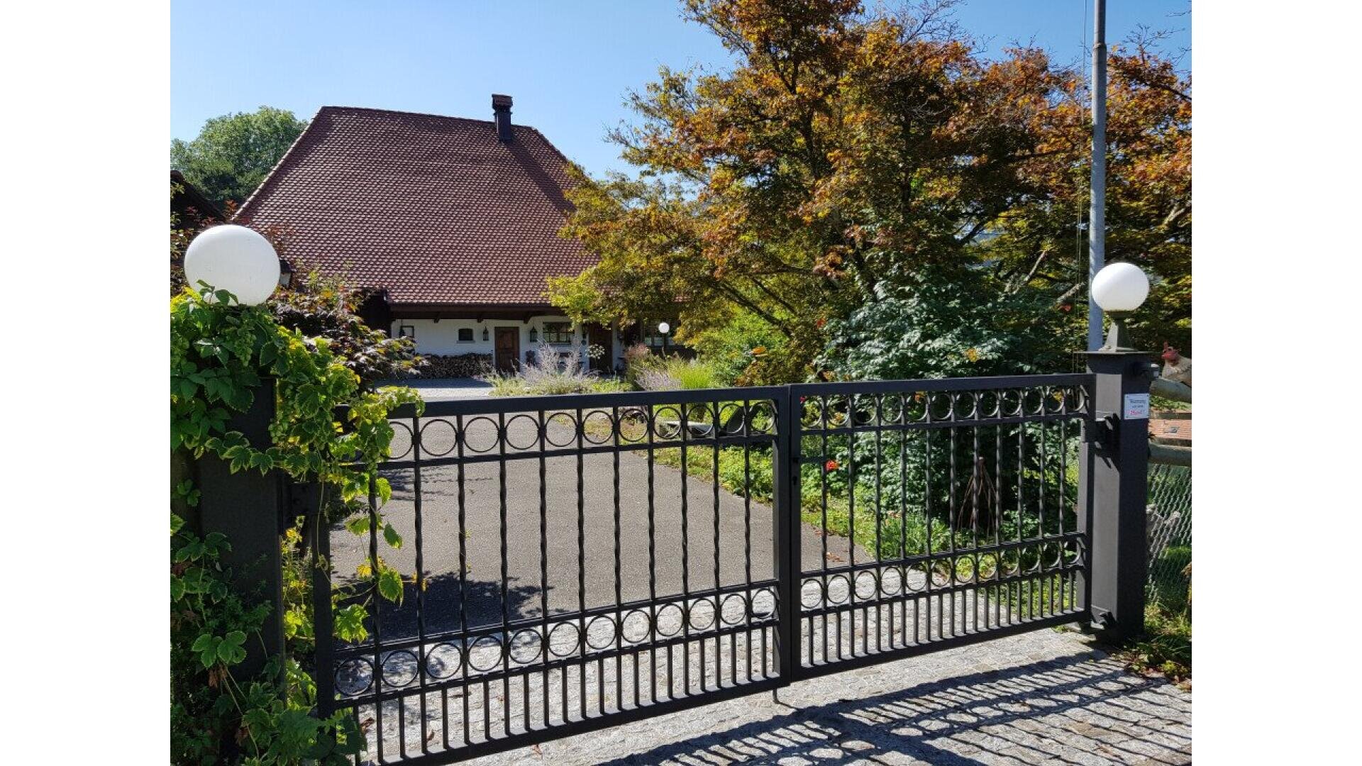 A black metal gate with circular designs stands closed in front of a driveway leading to a house with a red-tiled roof, surrounded by trees and greenery on a sunny day.