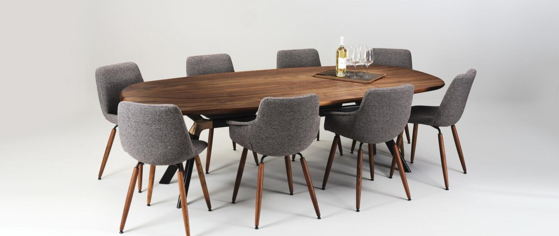 Ein moderner ovaler Esstisch aus Holz mit acht grau gepolsterten Stühlen. Auf dem Tisch steht ein Tablett mit einer Flasche Weißwein und vier Weingläsern. Der Hintergrund ist schlicht weiß.