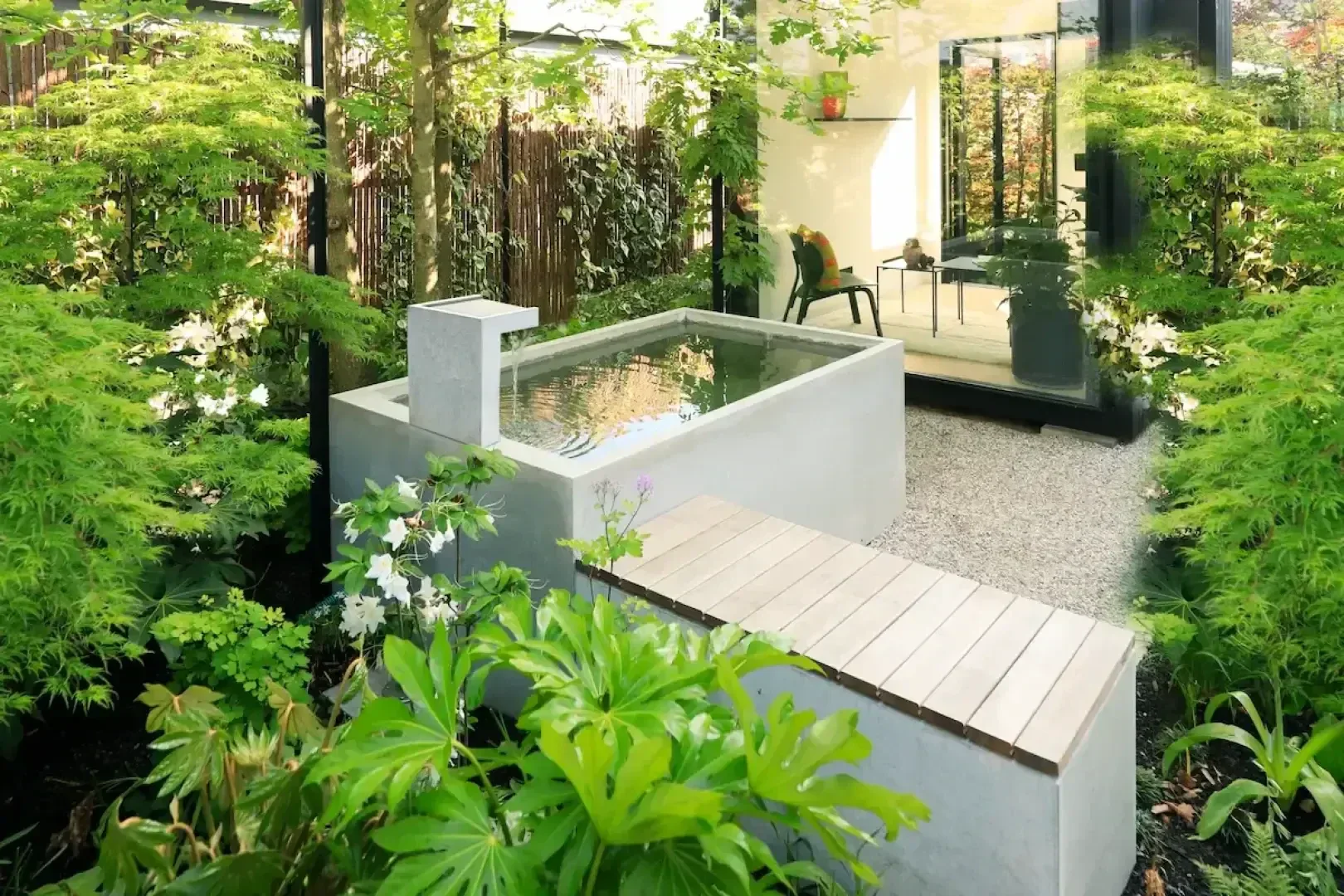 A modern garden features a small rectangular reflecting pool with a wooden bridge, surrounded by lush green plants and trees. A glass door opens to a sunlit indoor area with seating.