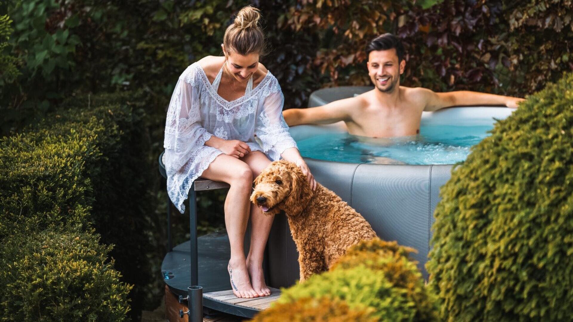 A woman in a white dress sits at the edge of a hot tub, petting a curly-haired dog. A man sits relaxed in the hot tub behind her. They are outdoors, surrounded by green hedges and plants.