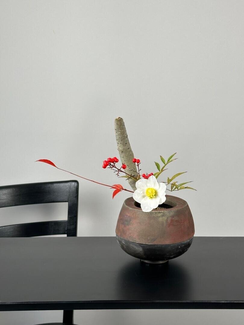 Ein minimalistisches Blumenarrangement in einer runden, rustikalen Vase steht auf einem schwarzen Tisch. Es besteht aus einer weißen Blume, roten Beeren und grünen und roten Blättern. Auf der linken Seite ist ein Teil eines schwarzen Stuhls vor einer schlichten grauen Wand zu sehen.