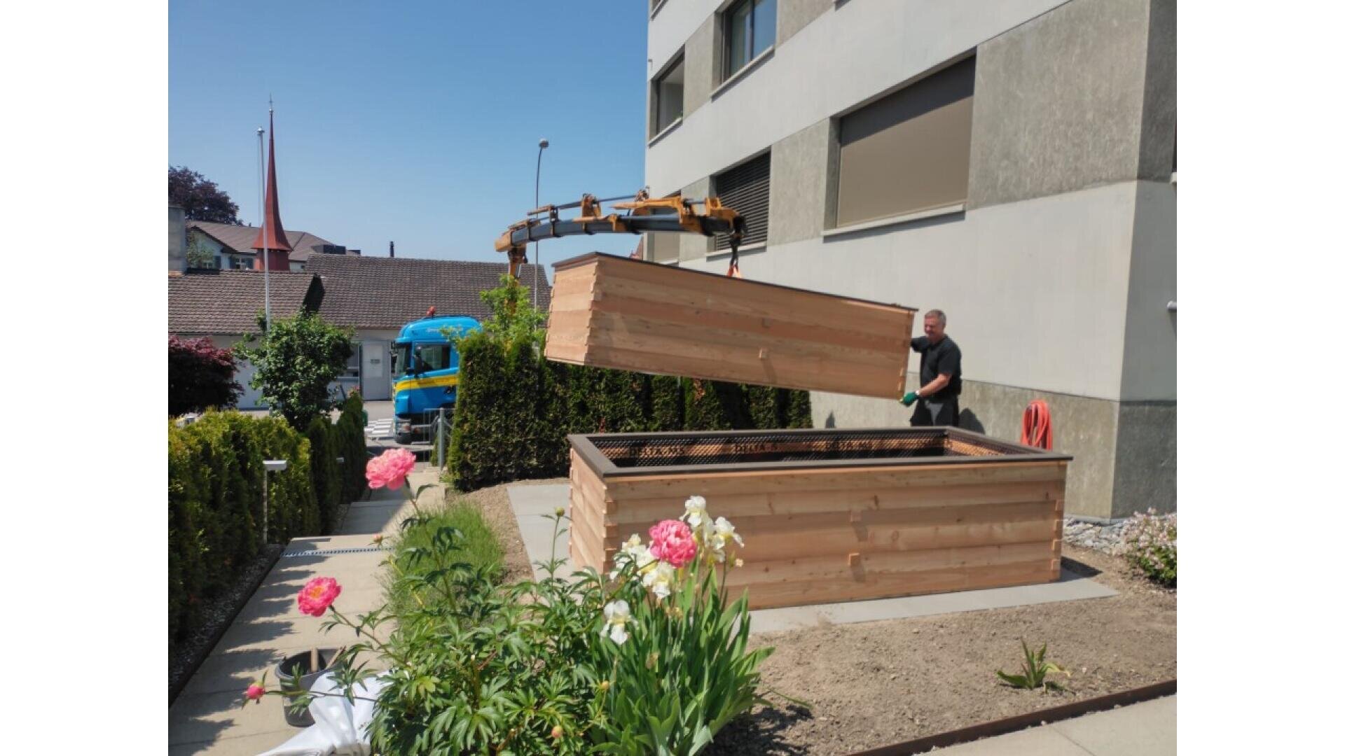 A person stands next to a large wooden planter box as a crane lifts its lid. The scene is outdoors by a building, with blooming pink flowers and a blue truck in the background.