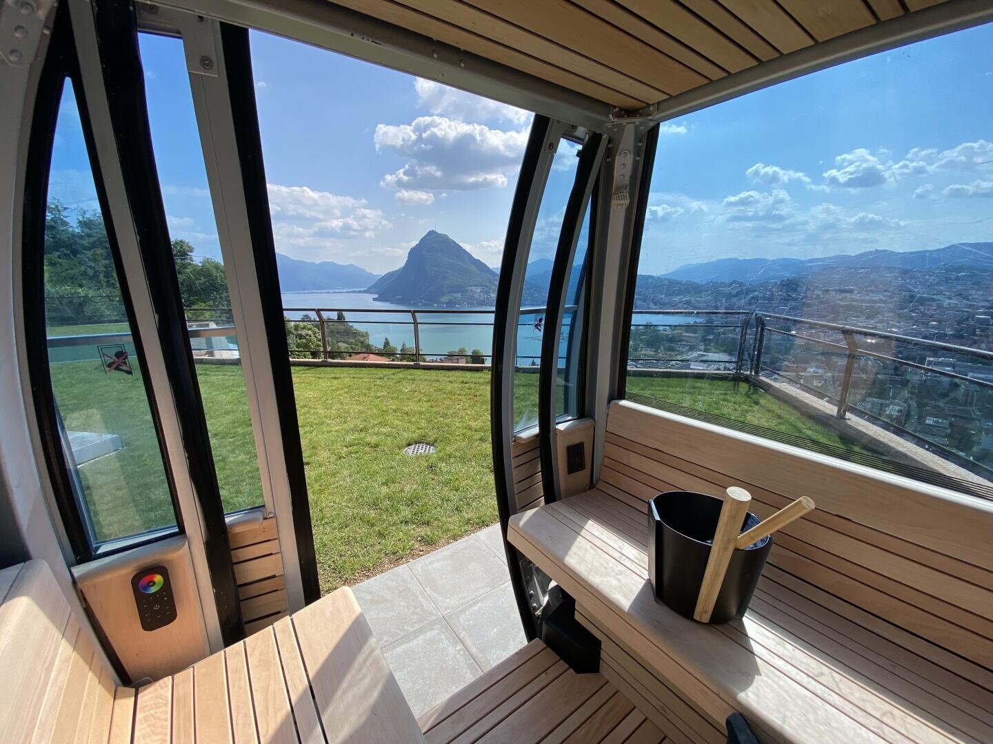 View from inside a modern wooden sauna with large windows, overlooking a green lawn, a lake, and mountains under a partly cloudy sky. A sauna bucket and ladle sit on a bench.