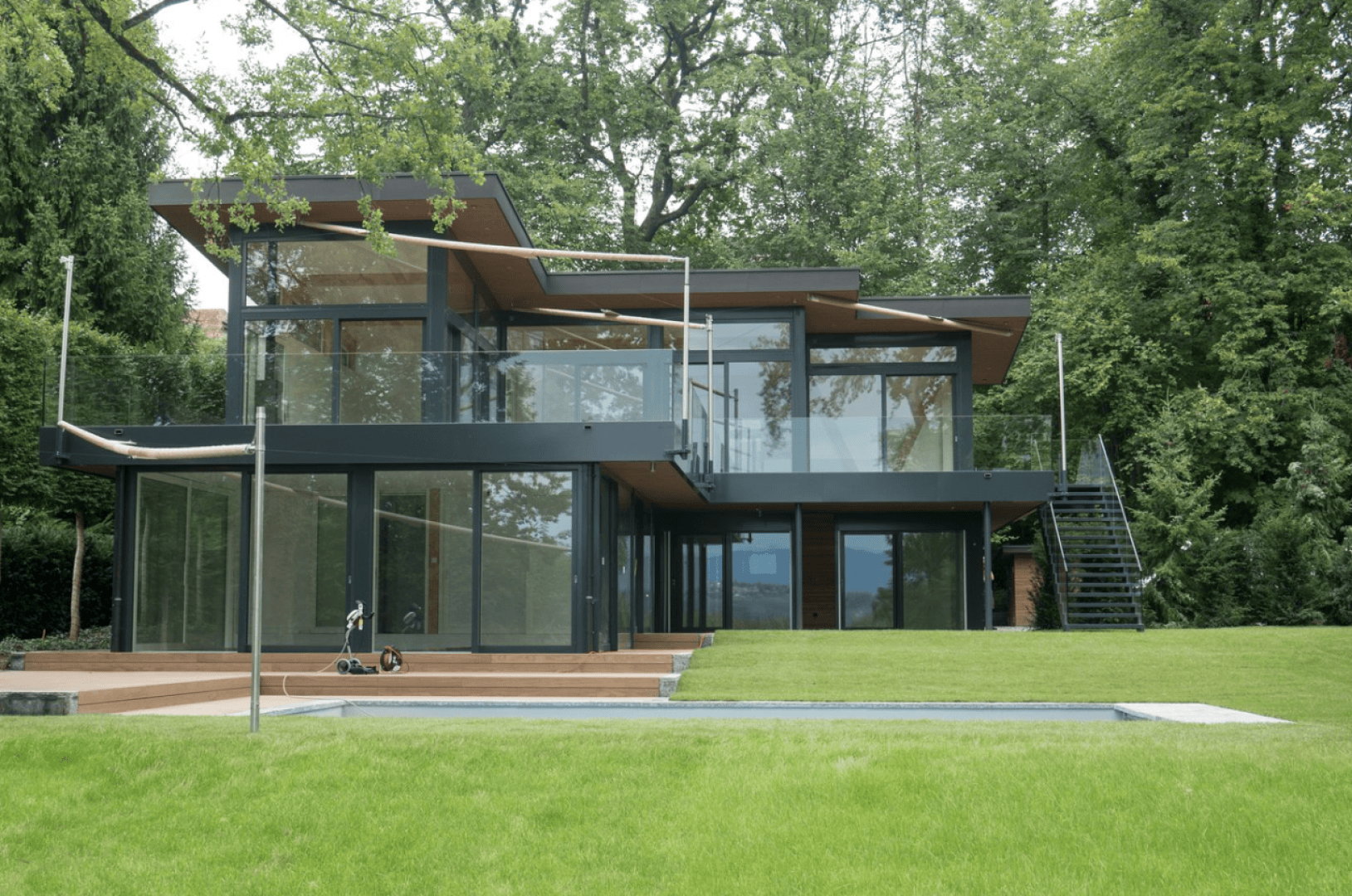 A modern two-story house with large glass windows and flat roofs, surrounded by lush green trees and grass, with a large lawn and a swimming pool in the front.
