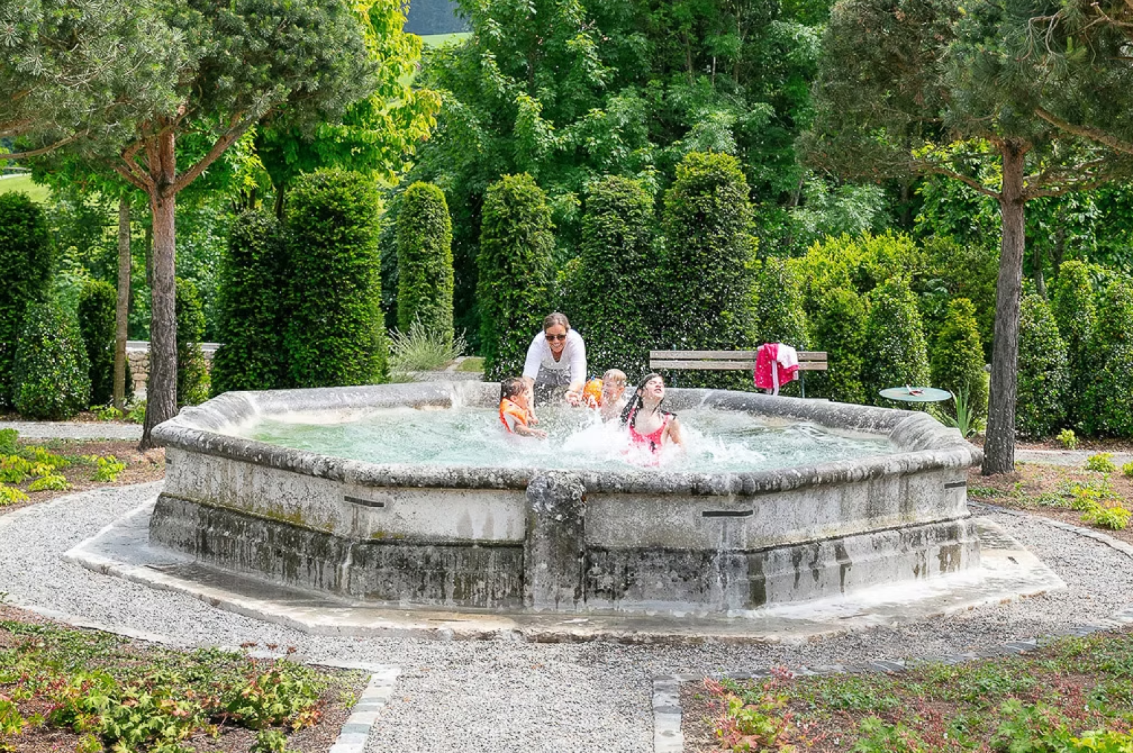 Vier Kinder und ein Erwachsener planschen und spielen an einem hellen, sonnigen Tag in einem großen, achteckigen Steinbrunnen, der von Grünflächen und Bäumen in einem Park umgeben ist.