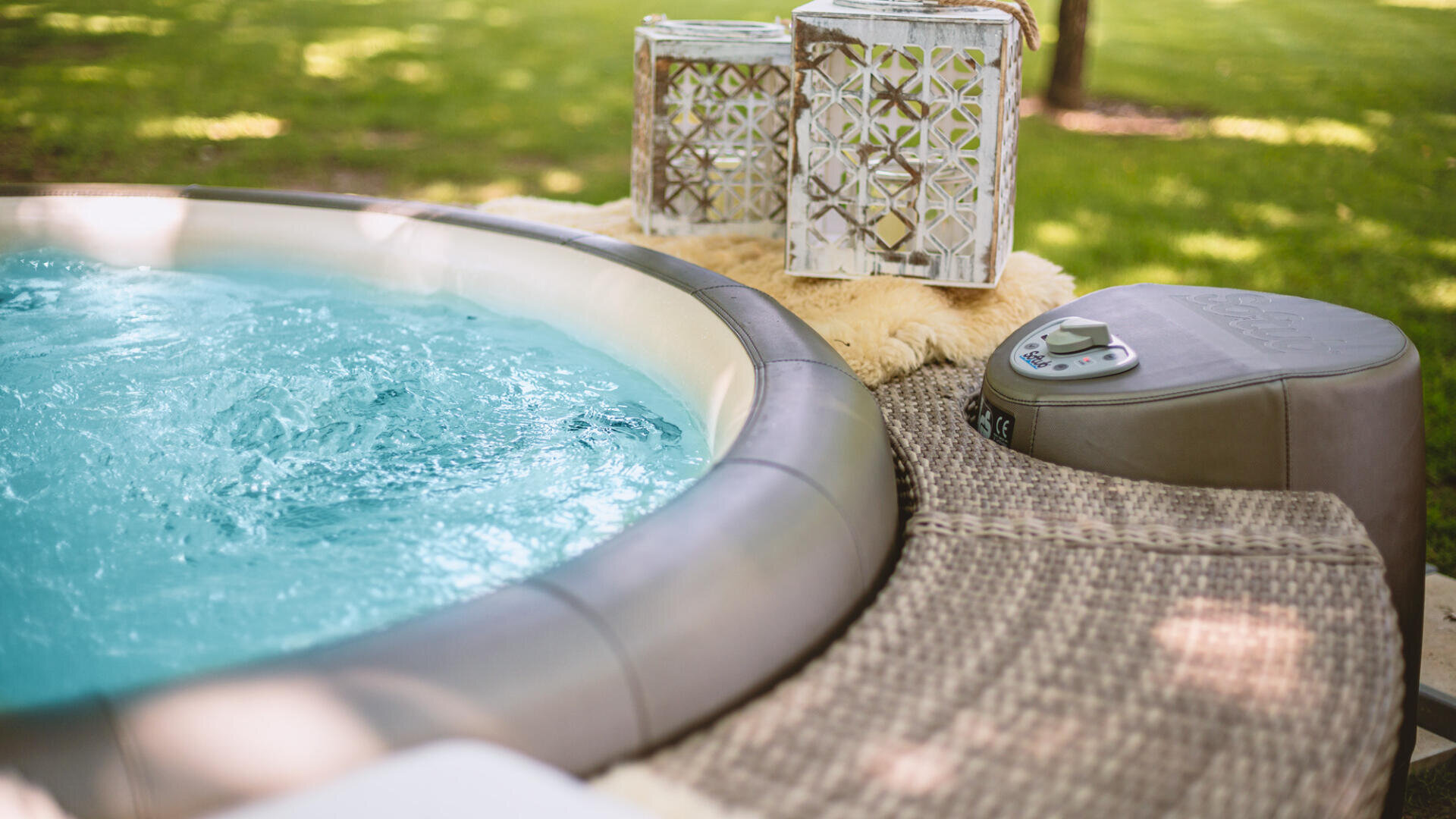 Nahaufnahme eines aufblasbaren Whirlpools im Freien mit sprudelndem Wasser, neben einer strukturierten Weidenfläche, auf der eine Steuereinheit und zwei dekorative weiße Laternen stehen. Grünes Gras und Bäume sind im Hintergrund verschwommen.