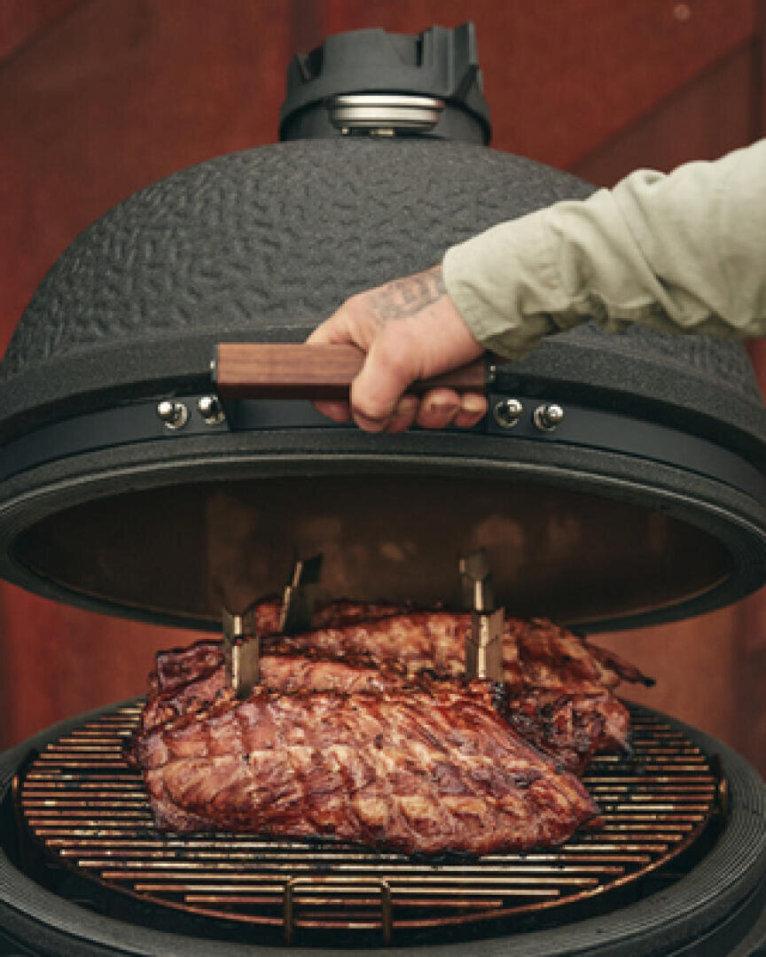 Eine Person hebt den Deckel eines runden, schwarzen Grills an und gibt den Blick auf die Rippen frei, die auf dem Rost gegrillt werden. Der Grill hat eine strukturierte Oberfläche und einen Holzgriff.