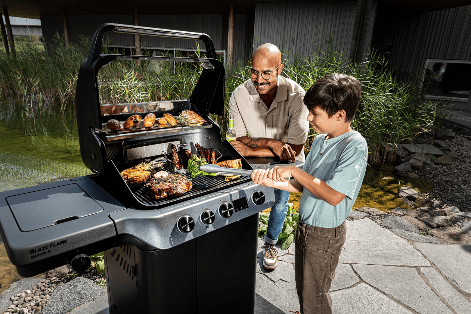 Ein Mann und ein Junge grillen verschiedene Fleisch- und Gemüsesorten auf einem schwarzen Grill im Freien neben einem Teich. Sie lächeln und genießen das gemeinsame Kochen an einem sonnigen Tag.