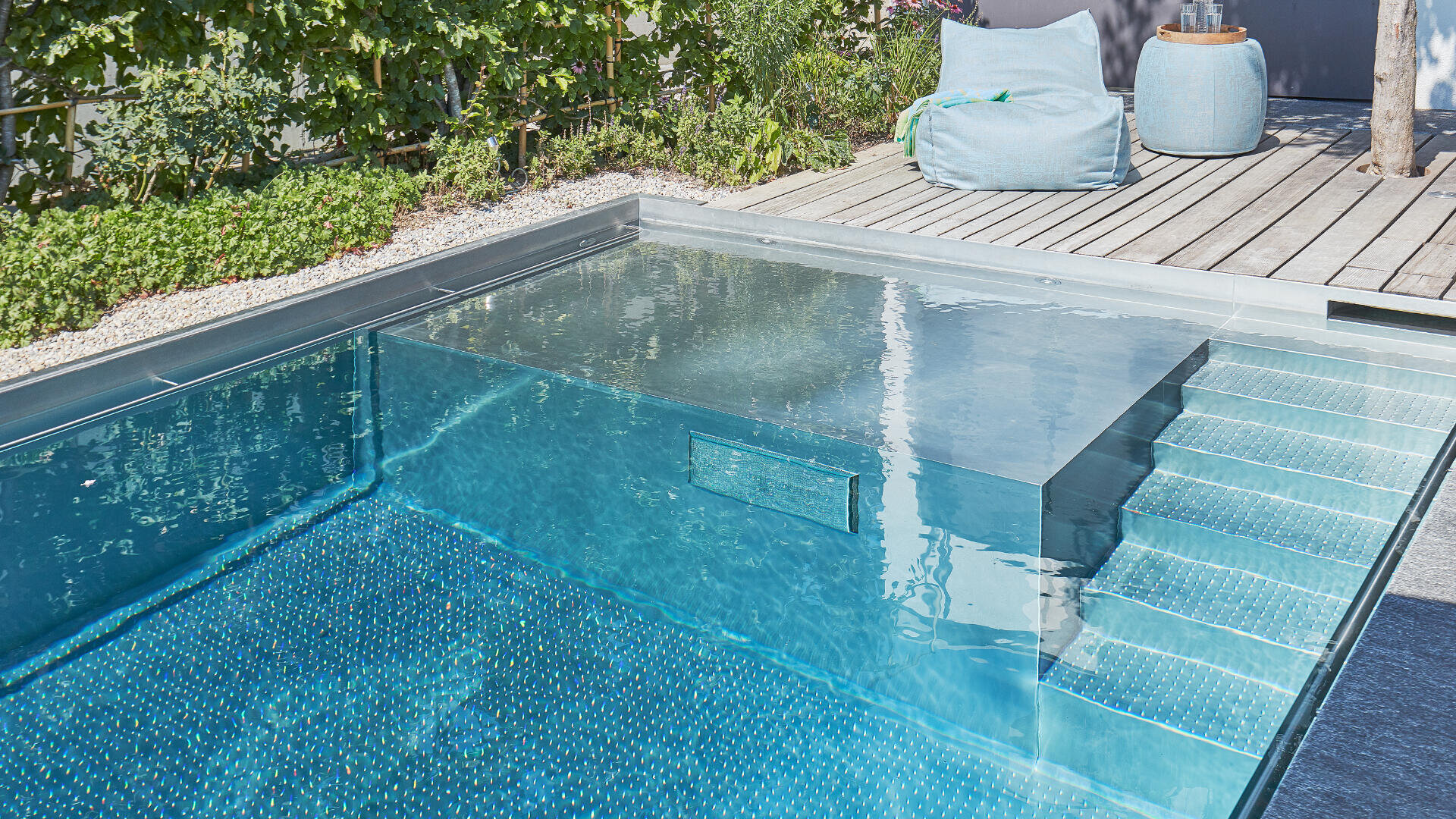 Modernes Außenschwimmbecken mit klarem, blauem Wasser, eingebauten Stufen und einem flachen Liegebereich. Der Pool befindet sich neben einer Holzterrasse mit Grünpflanzen, einem großen blauen Sitzsack und einem runden Tisch.