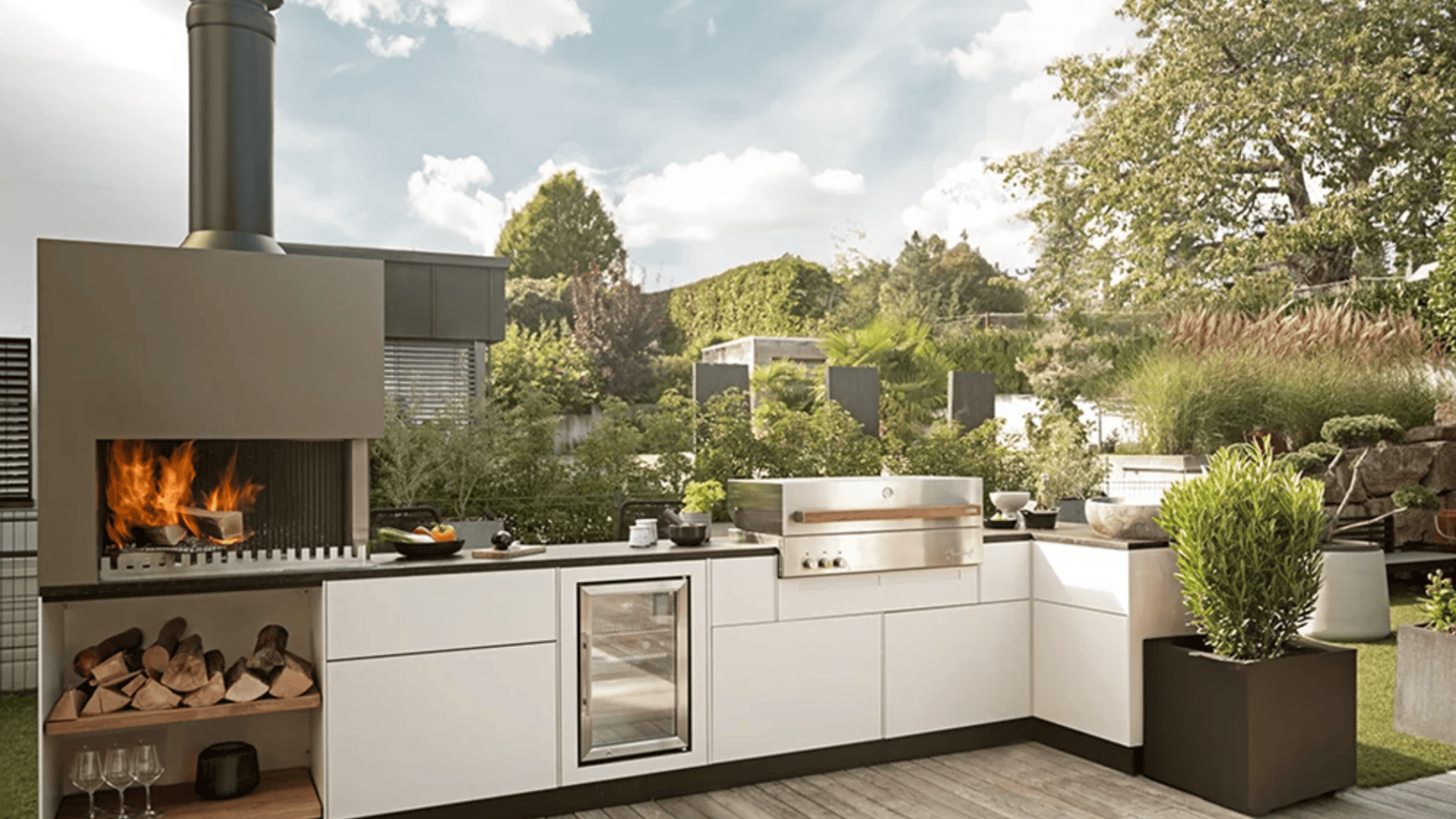 Moderne Außenküche mit eingebautem Grill, Feuerstelle mit brennendem Holz, Arbeitsplatte, Spüle, kleinem Kühlschrank und Topfpflanzen auf einer von Grün umgebenen Terrasse unter einem teilweise bewölkten Himmel.