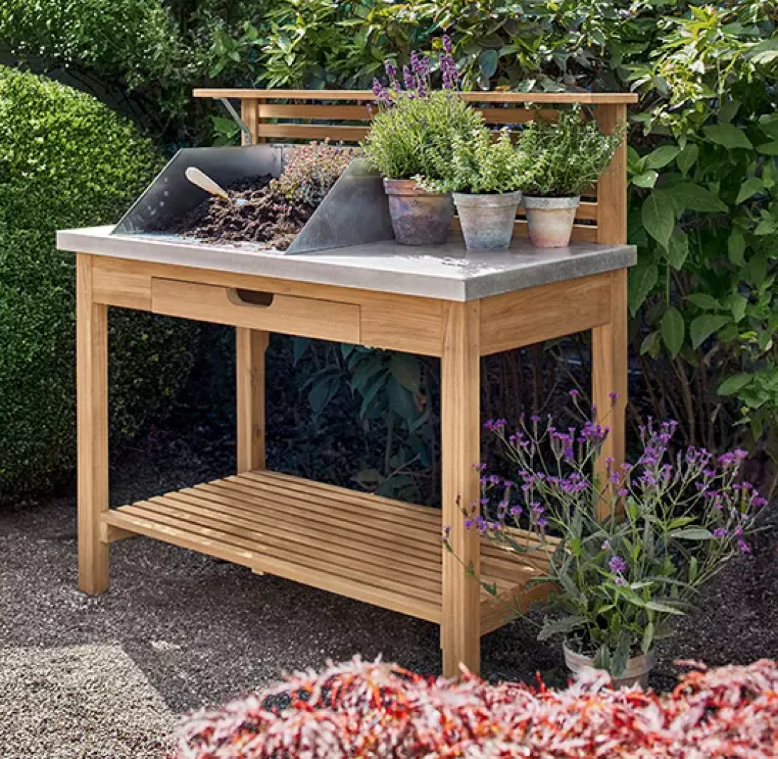 In einem Garten, umgeben von grünen Sträuchern und lilafarbenen Blumen, steht eine hölzerne Topfbank mit einer Metallschale, mehreren Kräutertöpfen und einer Kelle.