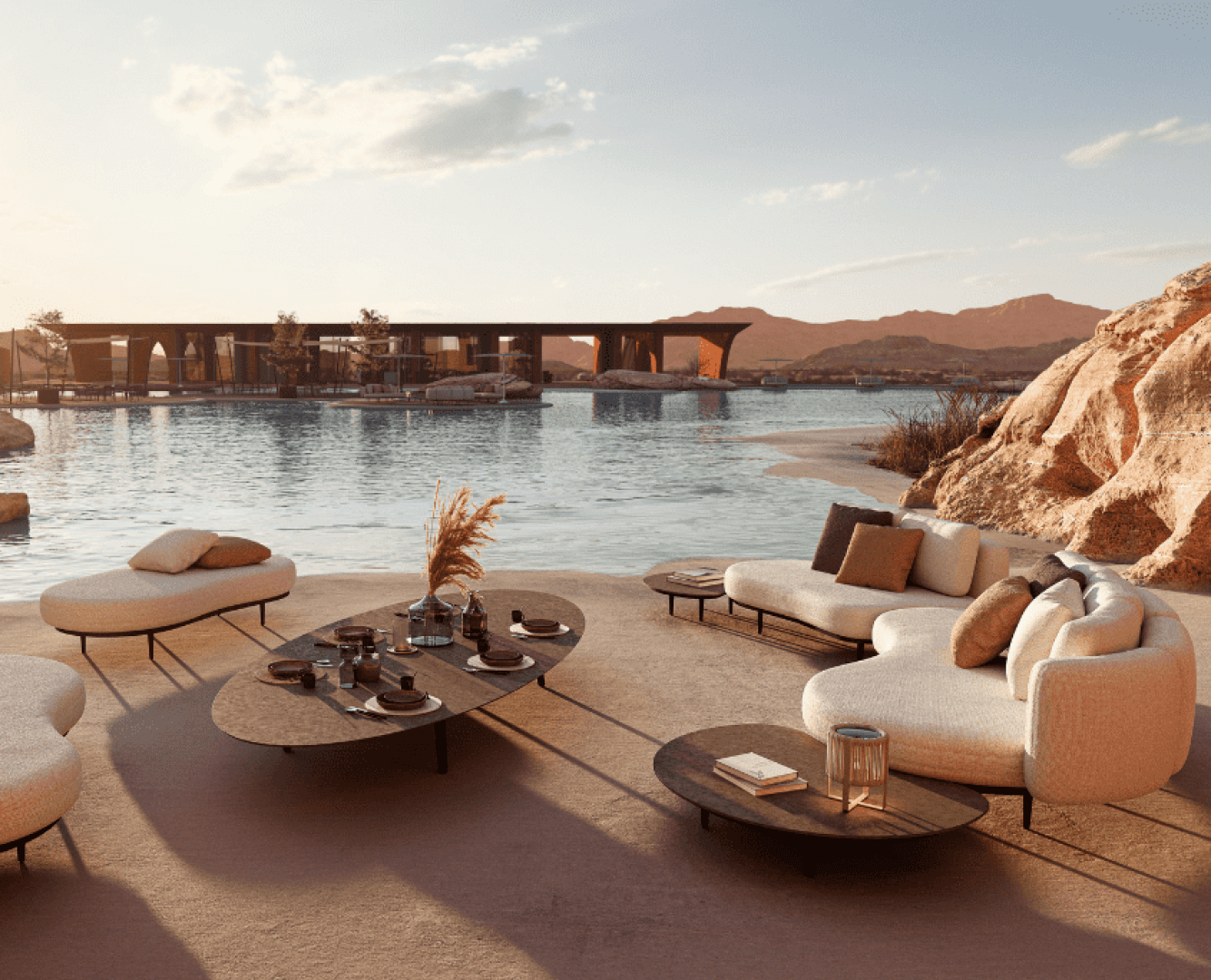 Moderner Lounge-Bereich im Freien mit beigen Sofas und niedrigen Tischen an einem ruhigen Wasserlauf, umgeben von felsiger Wüstenlandschaft und Bergen im Hintergrund bei Sonnenuntergang.