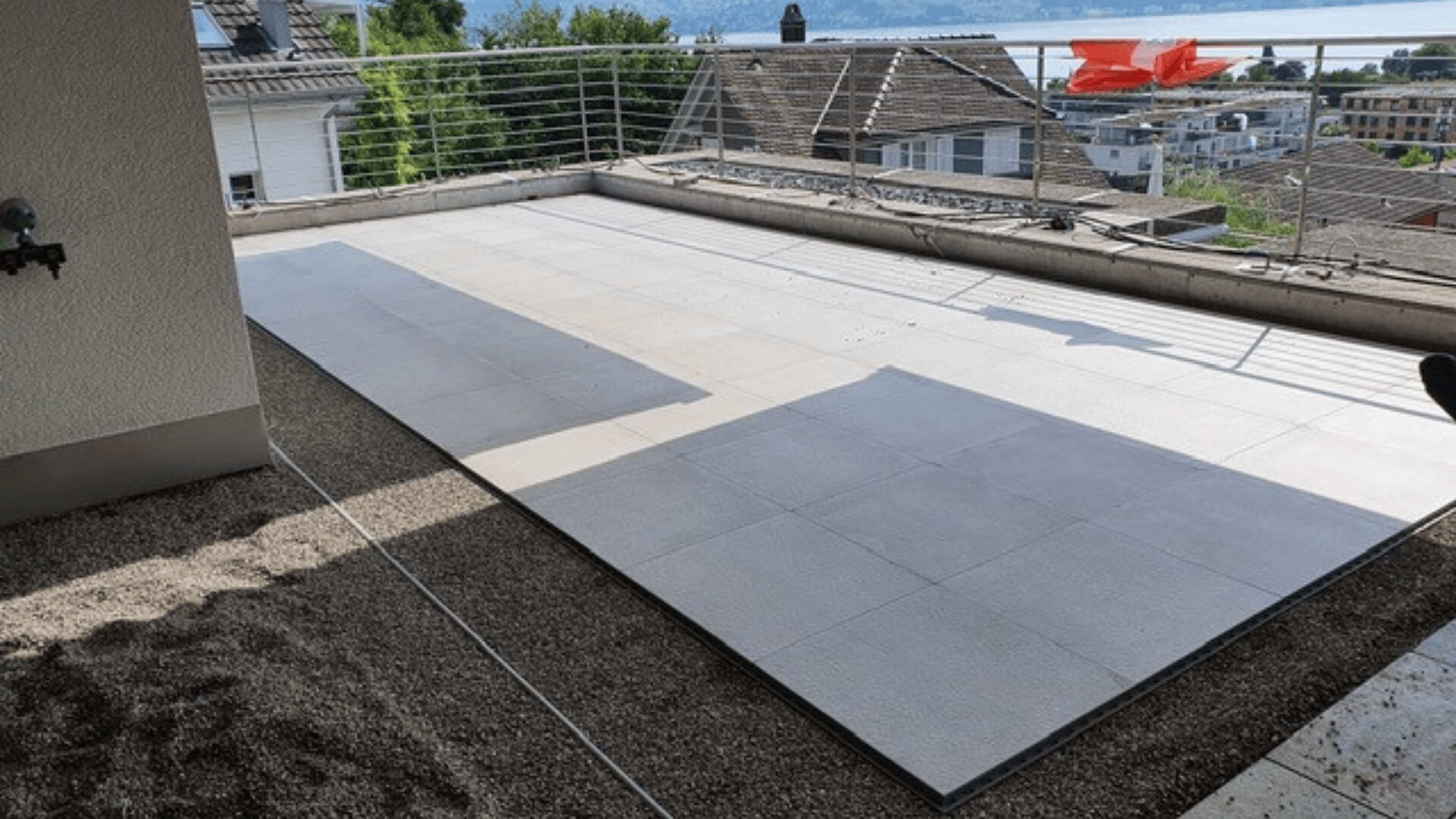 Eine Dachterrasse mit großen hellen Fliesen und einem Metallgeländer bietet einen Blick auf Häuser, Bäume und ein Gewässer. Um den gefliesten Bereich herum ist etwas Kies zu sehen.