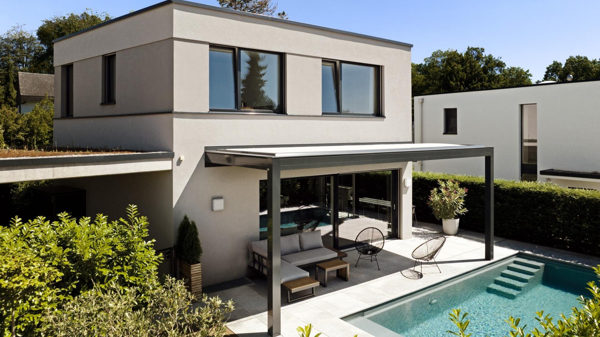 Modernes zweistöckiges Haus mit großen Fenstern, einer überdachten Terrasse mit Sitzgelegenheiten im Freien und einem Swimmingpool, umgeben von ordentlich gestutzten Hecken und Grünpflanzen unter einem klaren Himmel.