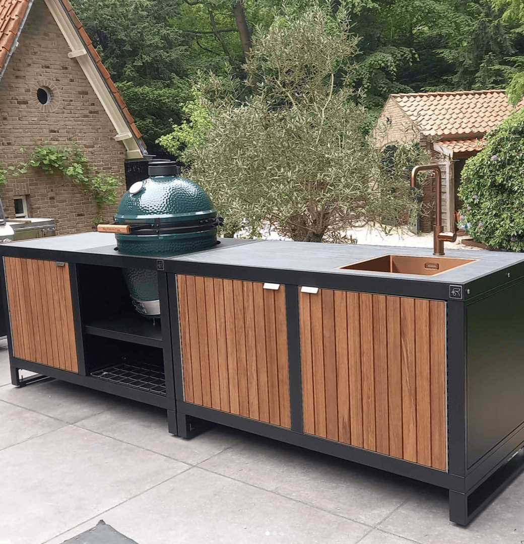 Outdoor kitchen with a wood and metal island, green ceramic grill, copper sink with tall faucet and storage cabinets, on a patio with trees and brick buildings in the background.