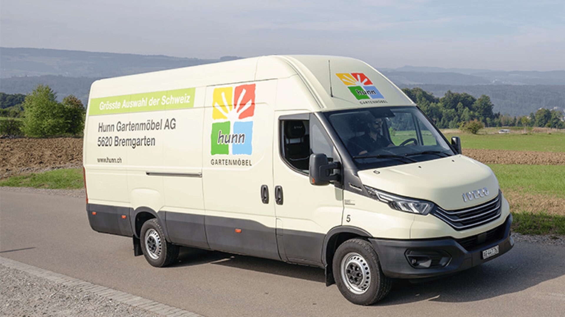 Ein weißer Lieferwagen mit dem Schriftzug Hunn Gartenmöbel und dem farbenfrohen Logo ist am Straßenrand neben Feldern geparkt, mit Hügeln und Bäumen im Hintergrund unter einem klaren Himmel.