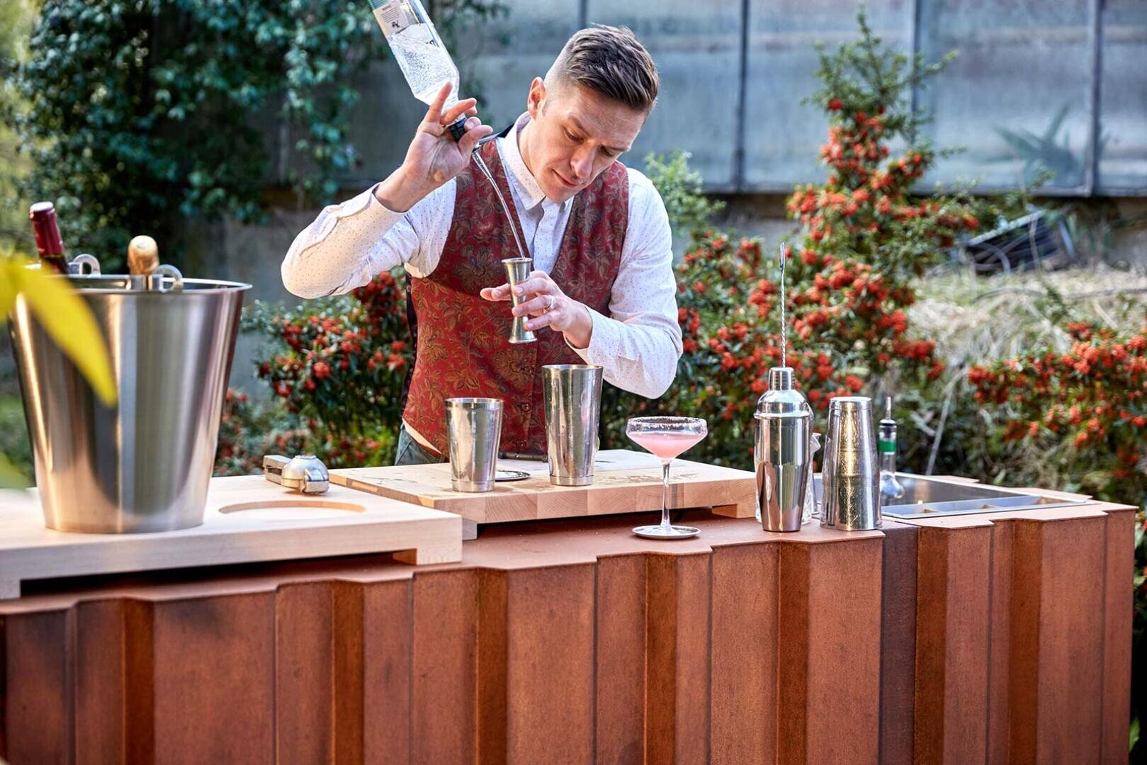A bartender wearing a patterned vest pours a clear liquid into a jigger behind an outdoor bar, surrounded by cocktail-making tools, a metal ice bucket, and a pink drink in a coupe glass. Shrubs and greenery are in the background.