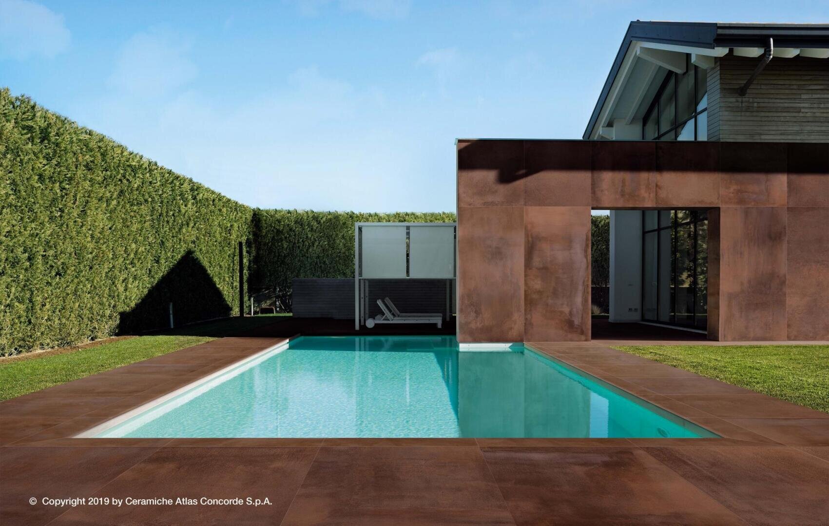 A modern house with large windows stands next to a rectangular swimming pool, surrounded by brown tiles and green hedges under a clear blue sky. Two sun loungers can be seen at the other end of the pool.