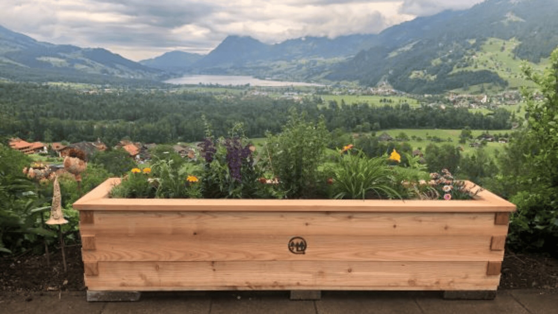 Ein hölzerner Pflanzkasten, gefüllt mit verschiedenen bunten Blumen und Grünpflanzen, steht auf einer Terrasse mit Blick auf ein malerisches Tal mit Bergen, Bäumen und einem See im Hintergrund unter einem bewölkten Himmel.