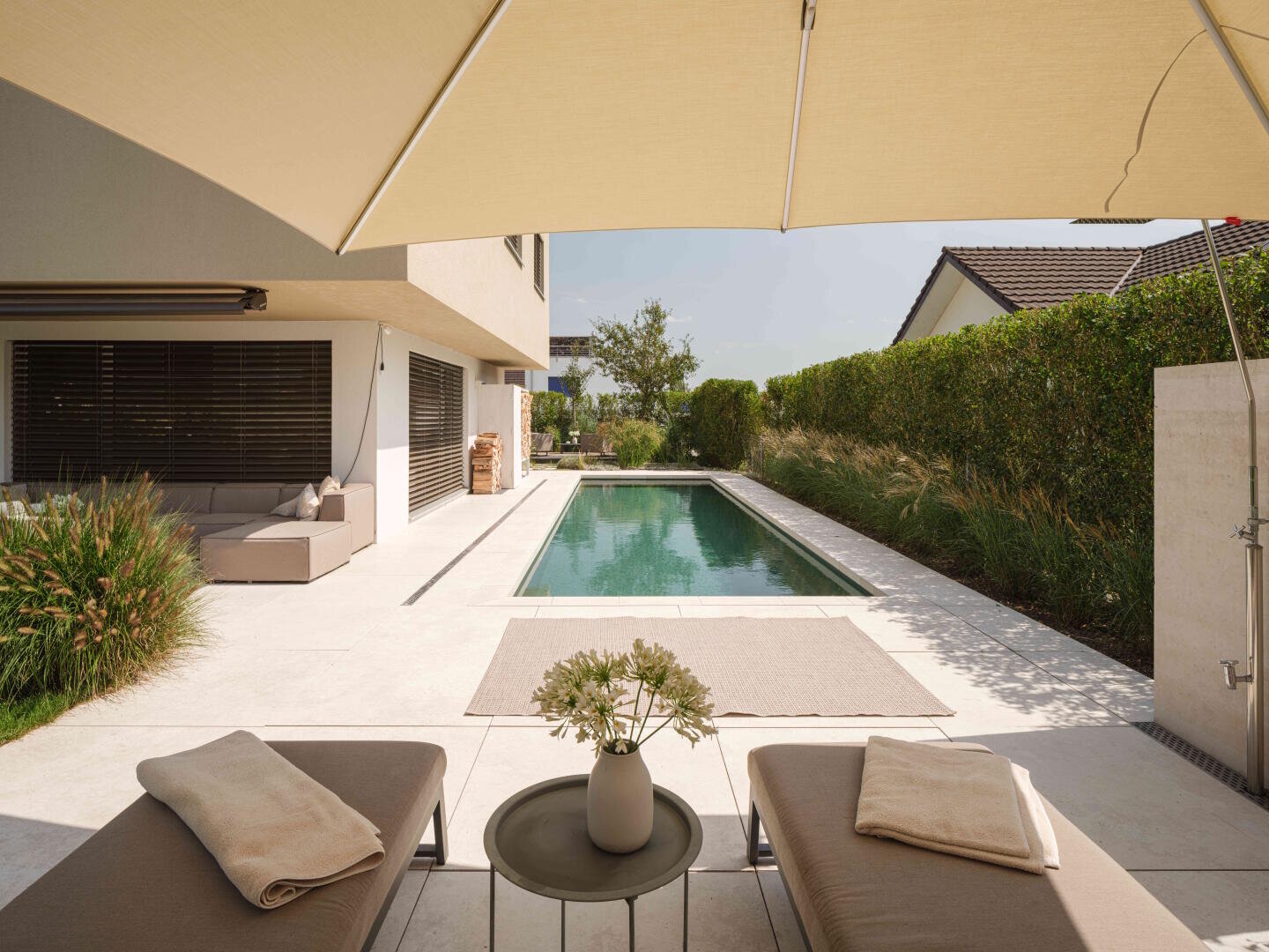 A modern backyard with a rectangular pool, two sun loungers with towels, a small table with flowers and a large beige parasol. A seating area and green areas line the edge of the pool.