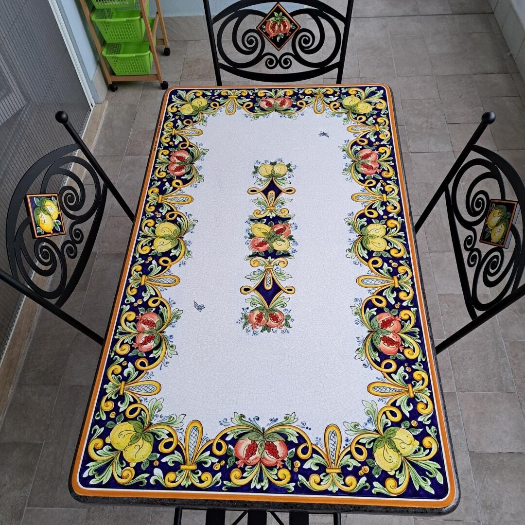A rectangular table with a colorful, ornate floral and fruit pattern, surrounded by three black metal chairs with decorative backs, set on a tiled floor indoors.