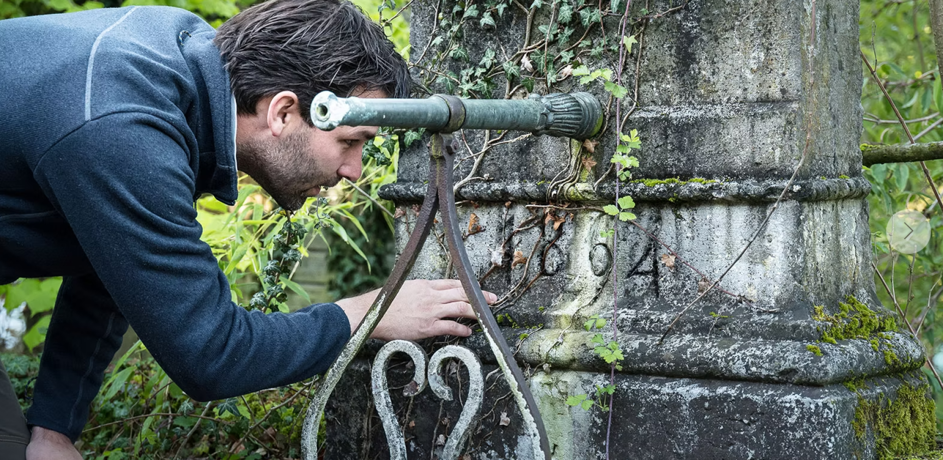 A man examines an old, ivy-covered stone building and looks closely at the numbers carved into the weathered surface.