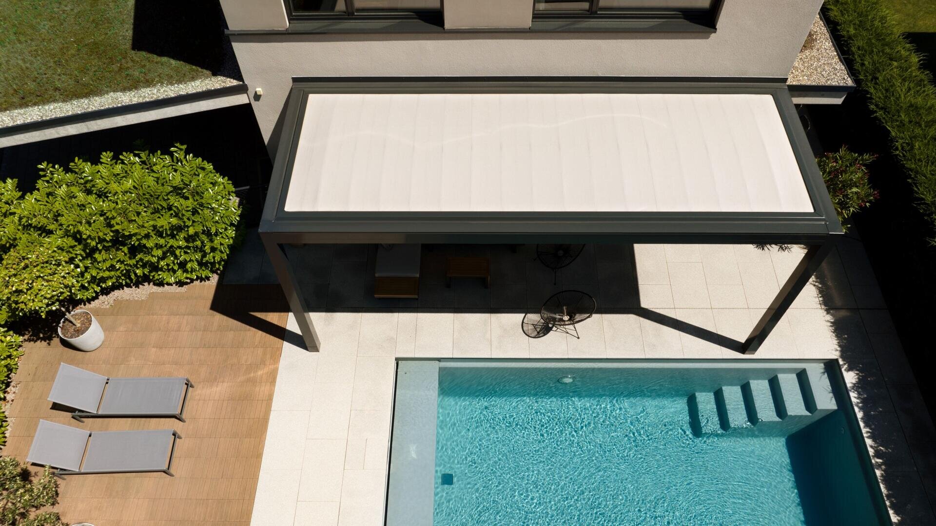 Luftaufnahme eines modernen Gartens mit einem Swimmingpool, einer von einer Pergola überdachten Terrasse am Pool, zwei Liegestühlen auf einer Holzterrasse und Grünpflanzen am Rande.